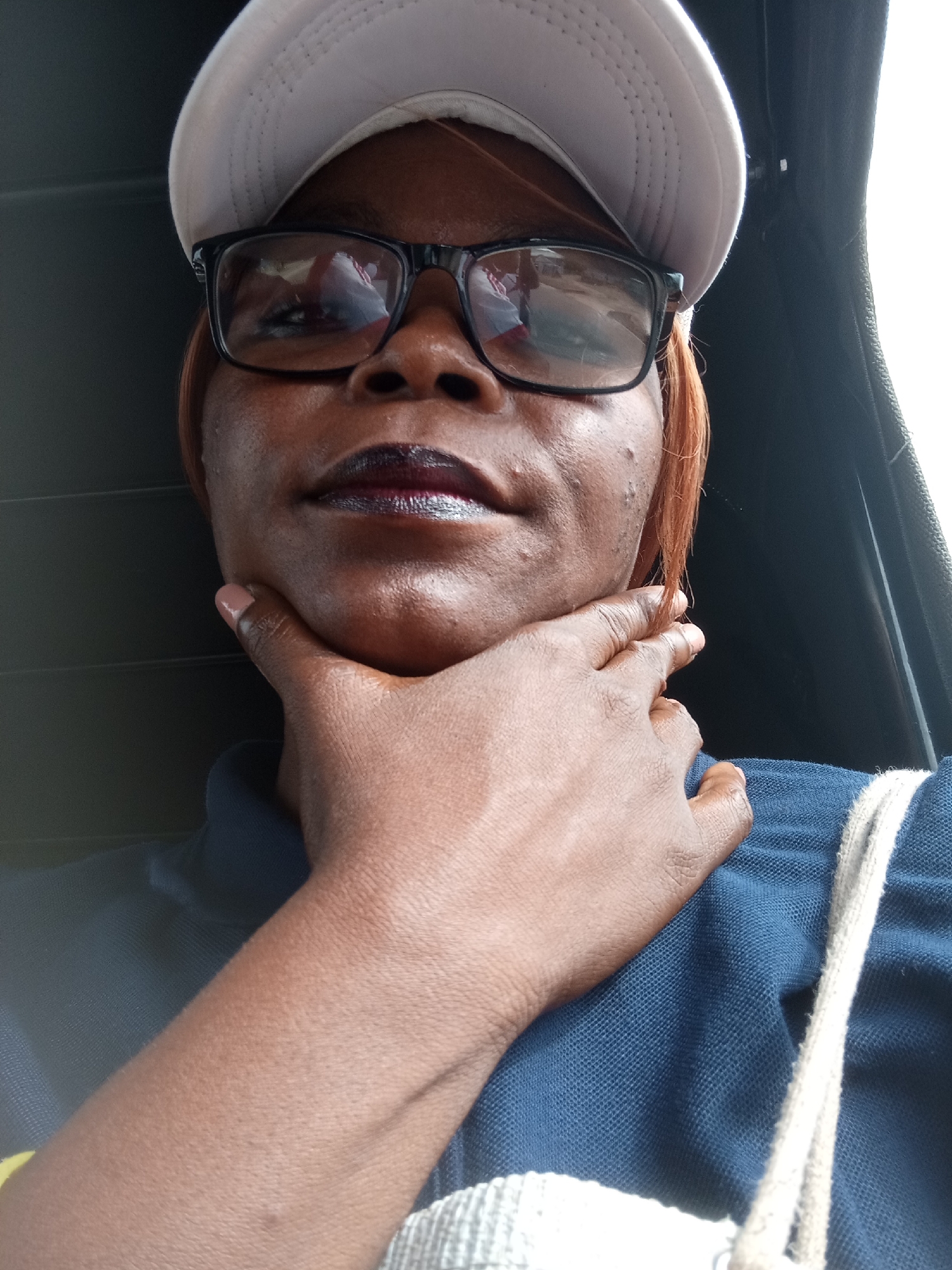 A medium shot captures a person from the chest up, facing forward with their head tilted slightly. They are wearing a light-colored baseball cap, dark-rimmed glasses, and a dark blue collared shirt. Their right hand is raised to their chin, with fingers resting on the jawline. The person has dark skin and dark hair visible from beneath the cap. The background is dark and appears to be the interior of a vehicle, with a bright white area to the right suggesting an open window or door. The lighting is natural and comes from the right side, casting shadows on the left. The image appears to have been taken outdoors, possibly in Zanzi, Nigeria, given the context.