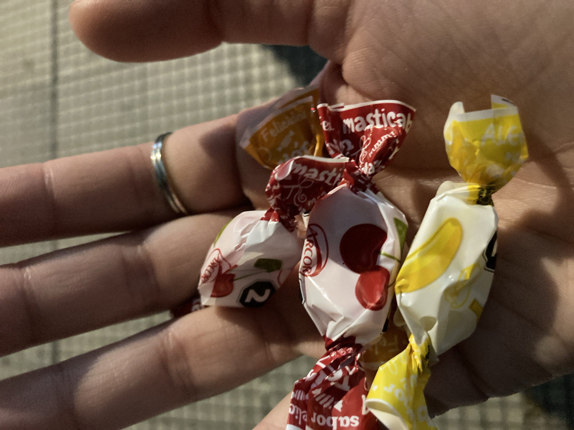 A close-up image captures a hand holding several individually wrapped candies. The hand, seen from the palm side, is gently cupped, showcasing the assortment of sweets. A simple silver ring adorns one of the fingers.
The candies are diverse in color and design. Some are wrapped in red foil, bearing the text "masticable" (chewable) and the "Arcor" brand logo, with "sabor" (flavor) also visible. Others are in white wrappers, featuring vivid red cherry illustrations and the "Arcor" logo, suggesting a cherry flavor. A couple of candies are wrapped in bright yellow foil; one clearly displays the word "Felicidad" (Happiness), and another shows "Ale" alongside a banana graphic, indicating a banana flavor.
The background is softly blurred, showing a light-colored, possibly patterned or tiled surface, providing little specific information about the location or time of day. The lighting is soft and even, highlighting the textures of the hand and the crinkled candy wrappers. The overall impression is of a casual moment, perhaps offering or receiving the candies.