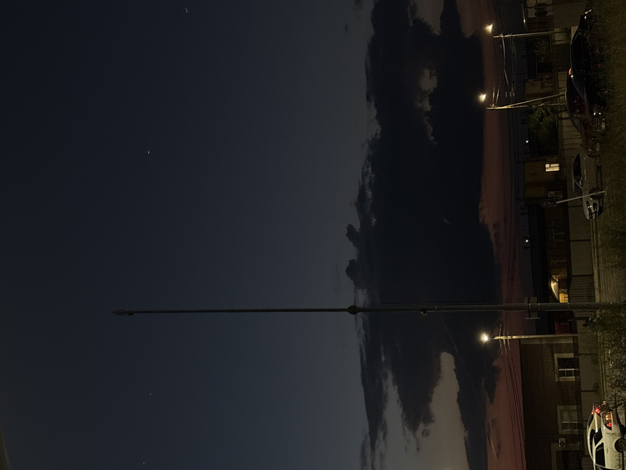 The image depicts a nighttime scene in Mikhaylovsk, Russia, likely during twilight or early evening. The sky is a deep, dark blue with some scattered clouds tinged with pink and orange, suggesting the last remnants of sunset or sunrise. Two small, bright pinpricks of light are visible in the sky, possibly stars or distant aircraft.
Dominating the foreground is a long, thin streetlight extending horizontally across the frame, with a bright light source illuminating a portion of the sky. Below and to the right, a street with buildings and parked vehicles is visible. One car, a white hatchback, is parked on the right side of the frame, with its taillights illuminated. Other vehicles are partially visible further down the street. The buildings appear to be residential or commercial, constructed with brick and featuring windows. Electrical wires crisscross the scene, strung between utility poles that are partially visible.
The overall mood is quiet and still, characteristic of a calm evening. The lighting suggests artificial illumination from streetlights and the car's taillights contrasting with the fading natural light in the sky. No people are visible in the image. There is no visible text.