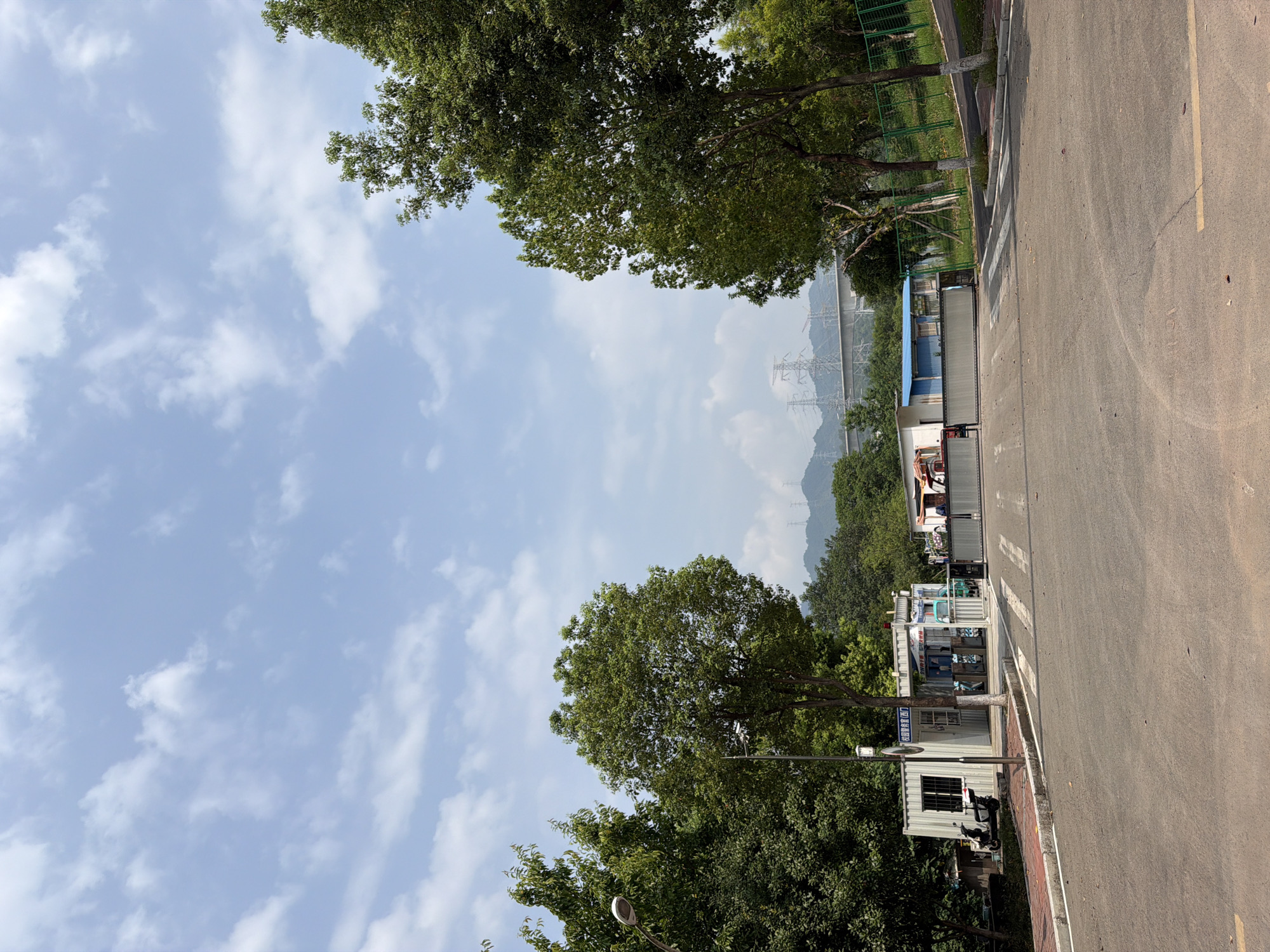 A wide shot captures an outdoor scene in Jiangjin, China, under a bright, partly cloudy sky. In the foreground, a paved road curves gently from left to right, with faint yellow lines marking its edges. A white pedestrian crossing is visible on the road.
Dominating the middle ground is a gated entrance to what appears to be a facility or park. The gate is closed, a long, metallic barrier. To the left of the gate is a small, white modular building with windows and a sign, serving as a security booth. A scooter is parked to the left of this building. To the right of the modular building and before the gate, a blue building with a white trim is situated. Several other structures, possibly temporary housing or storage units, are visible behind the gate and to the right.
Lush green trees line both sides of the road and the area surrounding the buildings, with some taller trees framing the right side of the frame. In the background, rolling hills are visible, and prominent electrical transmission towers and lines extend across the landscape, suggesting proximity to power infrastructure. A distant bridge can also be seen. The overall impression is a functional, possibly industrial or administrative area surrounded by greenery.