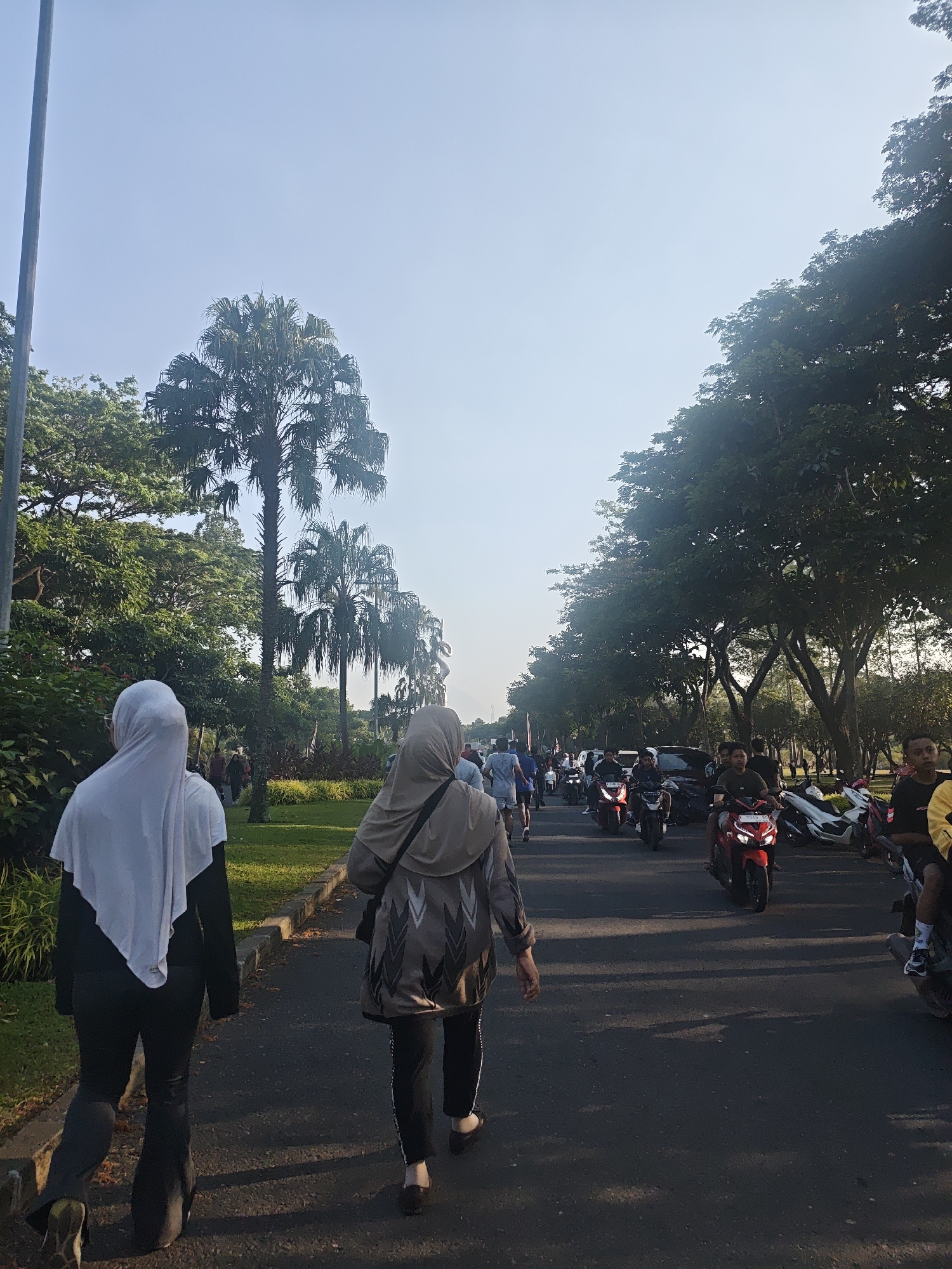 This image depicts a scene on a paved road lined with trees and a grassy median, likely in the city of Bacin, Indonesia. Several people are visible, some walking and others on motorcycles. In the foreground, two individuals are walking away from the camera, both wearing headscarves and loose-fitting clothing. The person on the left is wearing a white headscarf and black attire, while the person on the right wears a beige headscarf and a patterned jacket with black pants. The road is populated with numerous motorcycles, some occupied by riders and others parked. The environment suggests a public outdoor space, possibly a park or a street in a residential area, characterized by lush green foliage and a clear sky. The lighting indicates it is daytime, with sunlight casting shadows on the road.