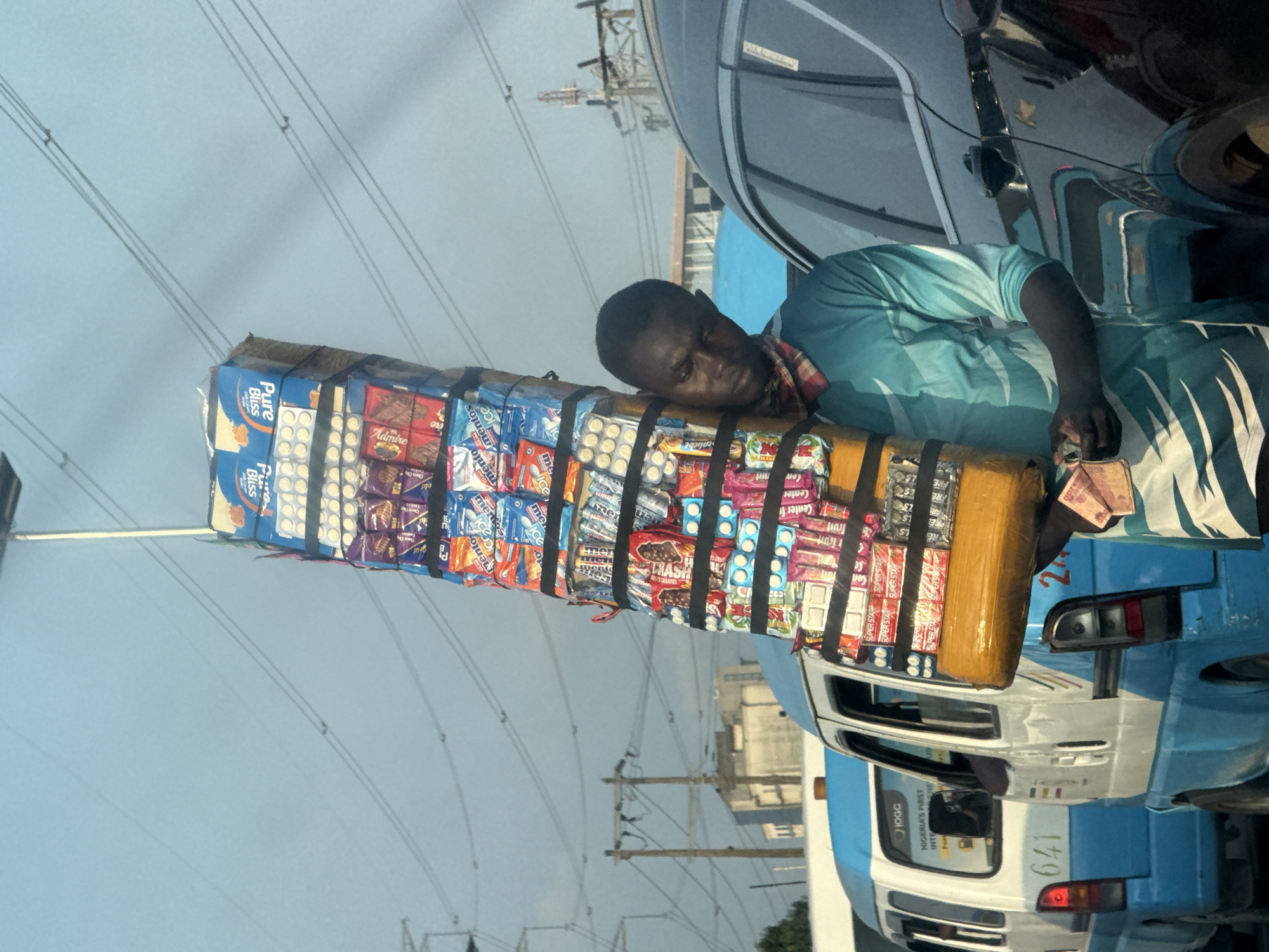 A young Black man is seen in Lekki, Nigeria, carrying a tall, stacked load of various snacks and confectioneries balanced on his head. He is wearing a blue and white tie-dye shirt and a checkered cloth around his neck. The man is holding some currency in his right hand. He is standing next to a dark-colored car and in front of two blue and white minibuses. The scene appears to be a busy street with electrical wires overhead. The lighting suggests it is daytime, possibly late afternoon, given the low angle of the sun and the shadows.