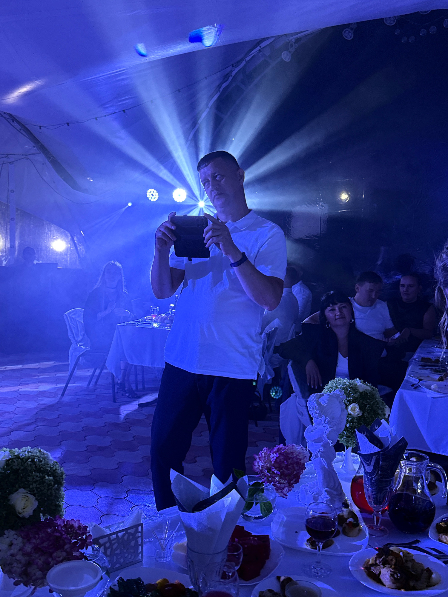 The image depicts an indoor event with blue lighting and theatrical spotlights illuminating the scene from above, creating dramatic light rays. In the foreground, a man in a white t-shirt and dark pants holds a tablet device, looking towards it. The room appears to be a banquet hall or a reception area, with tables set for dining. Several people are visible in the background, seated at tables. One woman in a dark jacket is seated at a table in the mid-ground, looking towards the camera. On the tables, there are floral arrangements, glassware, plates with food, and pitchers of drinks. The floor has a patterned tiled surface. The overall atmosphere suggests a social gathering or celebration. The location is stated as Пакли, Russia.