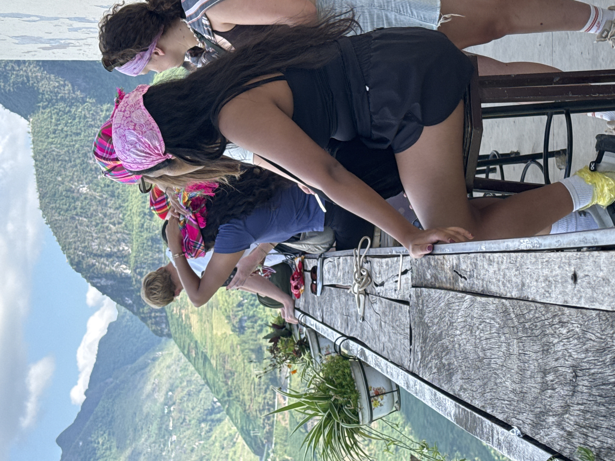 The image shows a group of people on an elevated wooden platform, likely a viewpoint, overlooking a mountainous landscape in Đồng Văn Commune, Vietnam. The landscape includes green, forested hills and a visible sky. Several individuals, mostly women, are present. One woman is kneeling on the platform’s edge, wearing black shorts and a tank top, with long, dark hair. Another person is wearing a pink patterned bandana. The platform is constructed of weathered wood, and there are potted plants along the edge. A metal railing runs along the edge of the platform.