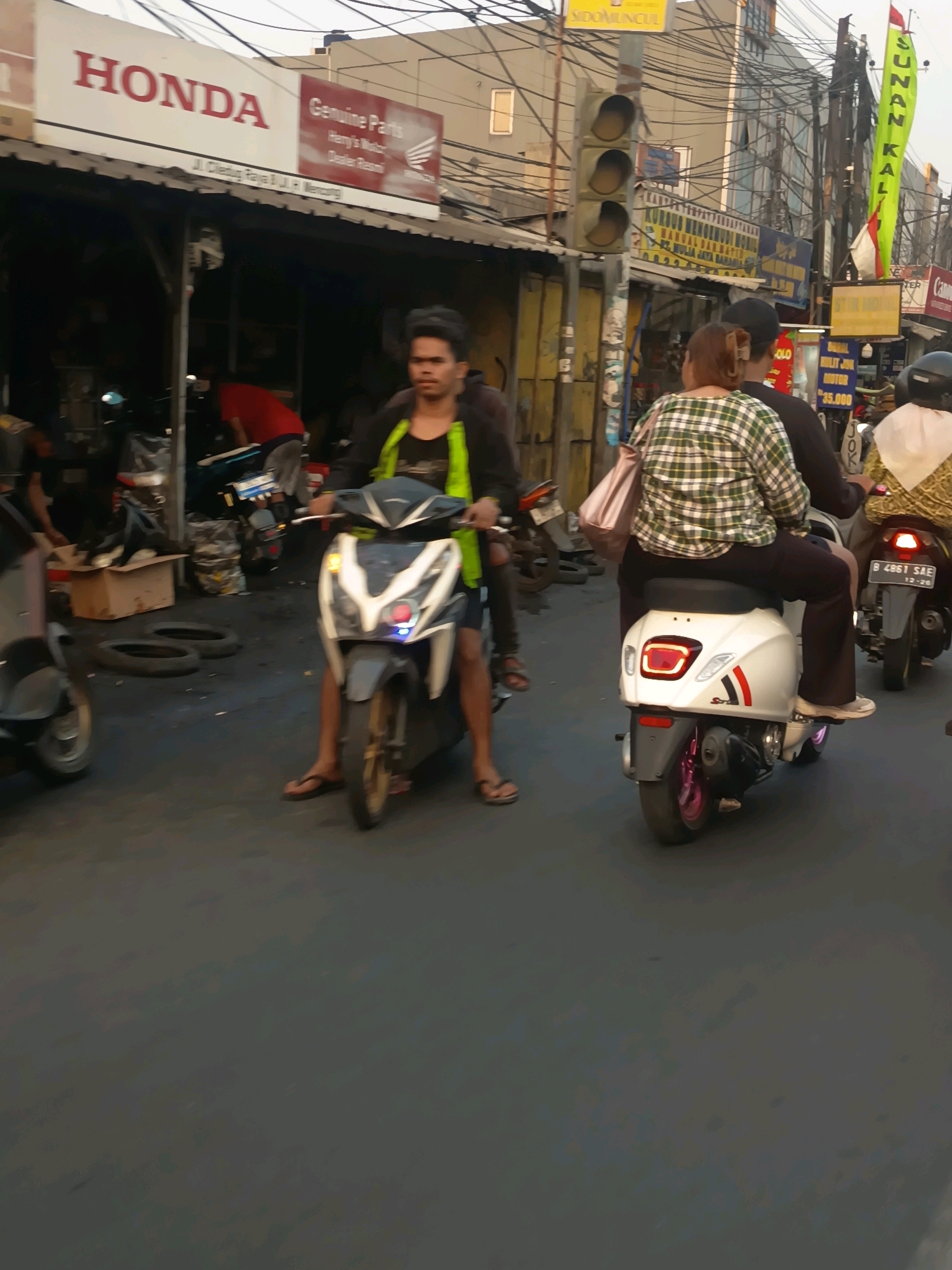 In Tangerang, Indonesia, a street scene features several individuals on motorcycles. In the foreground, a man wearing a green and black jacket and flip-flops rides a white scooter. To his right, a woman in a green and white checkered shirt and dark pants is a passenger on a white scooter, carrying a pink bag. Behind them, another person on a scooter is partially visible. In the background, a Honda dealership sign is prominent, indicating the location as a commercial area. The street is paved, and various shops and signs are visible along the roadside, suggesting a bustling urban environment. Traffic lights and power lines are also present, characteristic of Indonesian city streets. The overall atmosphere appears to be that of a typical day in a busy Indonesian city.
