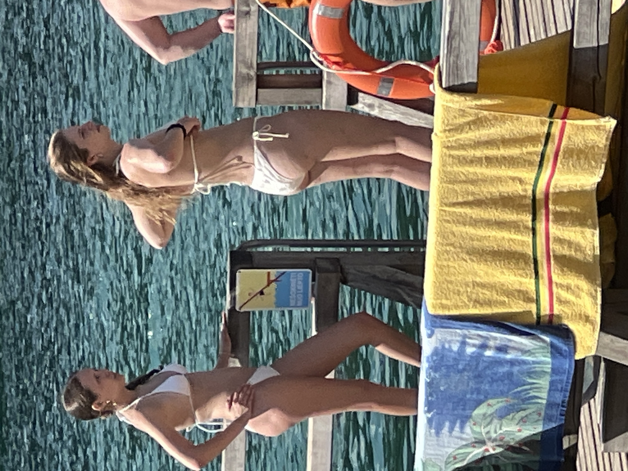 The image depicts two young women in bikinis standing near a body of water, possibly a lake or the sea. They are on a wooden pier, with towels draped over a railing in the foreground. One woman is facing left, wearing a white bikini and with her hair in a braid. The other woman is facing right, also in a white bikini, with her hair down. There is a life preserver ring visible on the right side of the image. The environment suggests a recreational setting on a sunny day. The location is identified as the city of Vilnius, Lithuania.