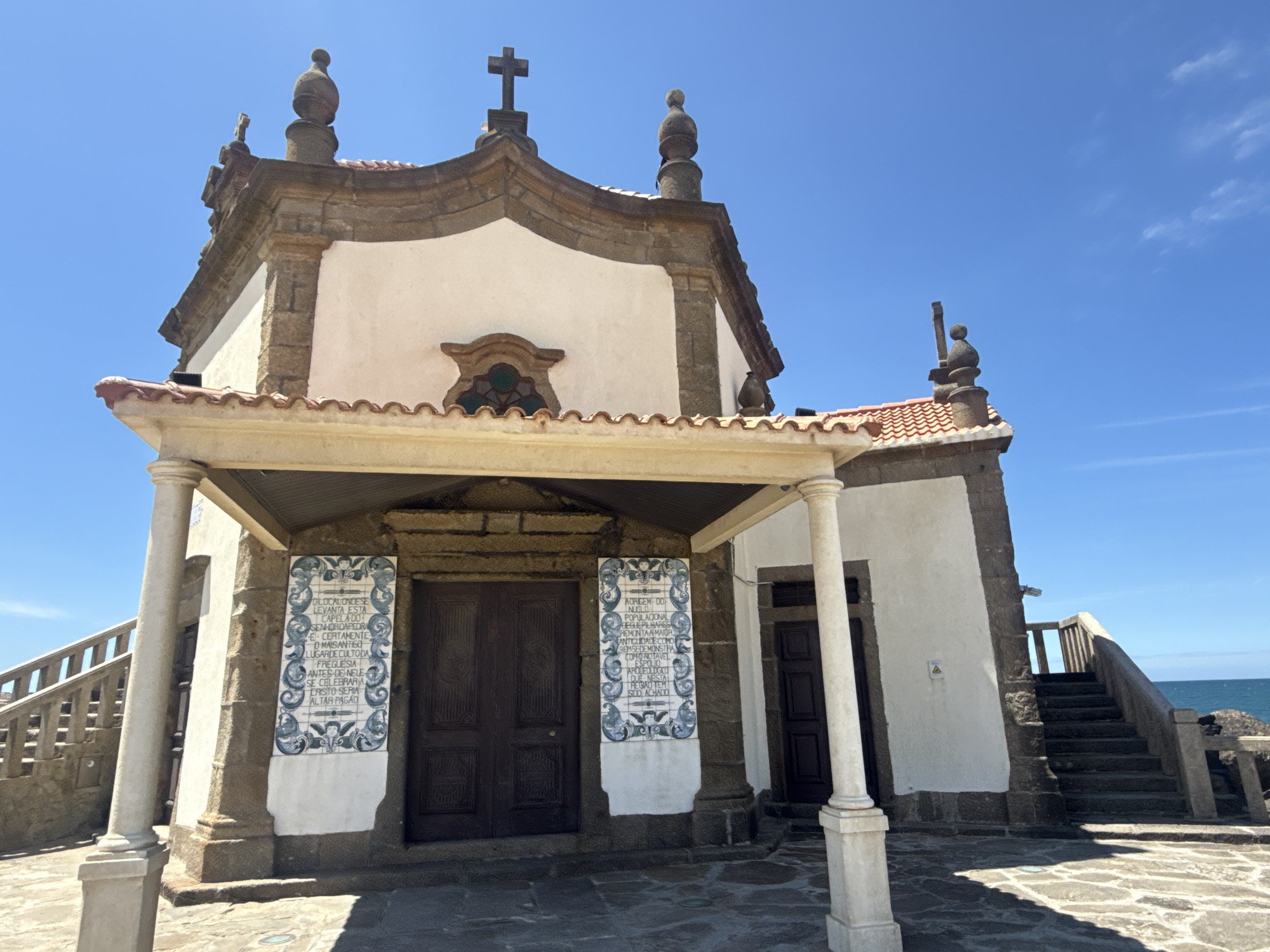 The image depicts a church building in Vila Nova de Gaia, Portugal. The church features a white facade, stone accents, and a red-tiled roof. A covered porch with columns provides shelter for the entrance. Two vertically oriented panels with white backgrounds and blue writing and decoration flank the main doors. A stone staircase is visible on the right side of the church, leading towards the ocean, which can be seen in the distance. The sky is bright blue with very few clouds. The church is likely a historical or religious landmark.