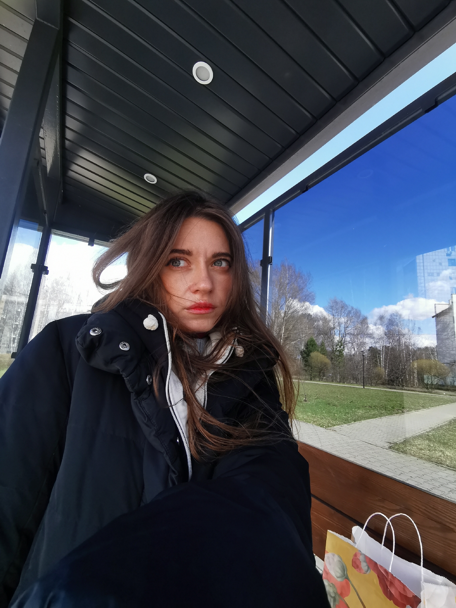 A young woman with long, wavy brown hair and light-colored eyes is depicted in a medium close-up, sitting inside a modern shelter. Her hair appears windswept. She wears a black puffer jacket over a white inner layer and has red lipstick. Her expression is pensive, with her gaze directed upwards and to the right, away from the viewer.
The setting appears to be a bus stop or similar public transport shelter. It features a dark, slatted ceiling with recessed circular lights and large glass panels forming one side. Through the glass, a clear, bright blue sky with scattered white clouds is visible. The outdoor scene includes a park-like area with green grass, bare trees, and a paved pathway. A modern building is partially visible in the distance to the right. The shelter also has a wooden bench where the woman is seated.
Beside her on the bench are two shopping bags: one yellow with an illustration of red poppy-like flowers and green leaves, and one plain white bag. The time of day is bright daylight, suggesting a clear day in late winter or early spring due to the bare trees. There is no legible text visible in the image.