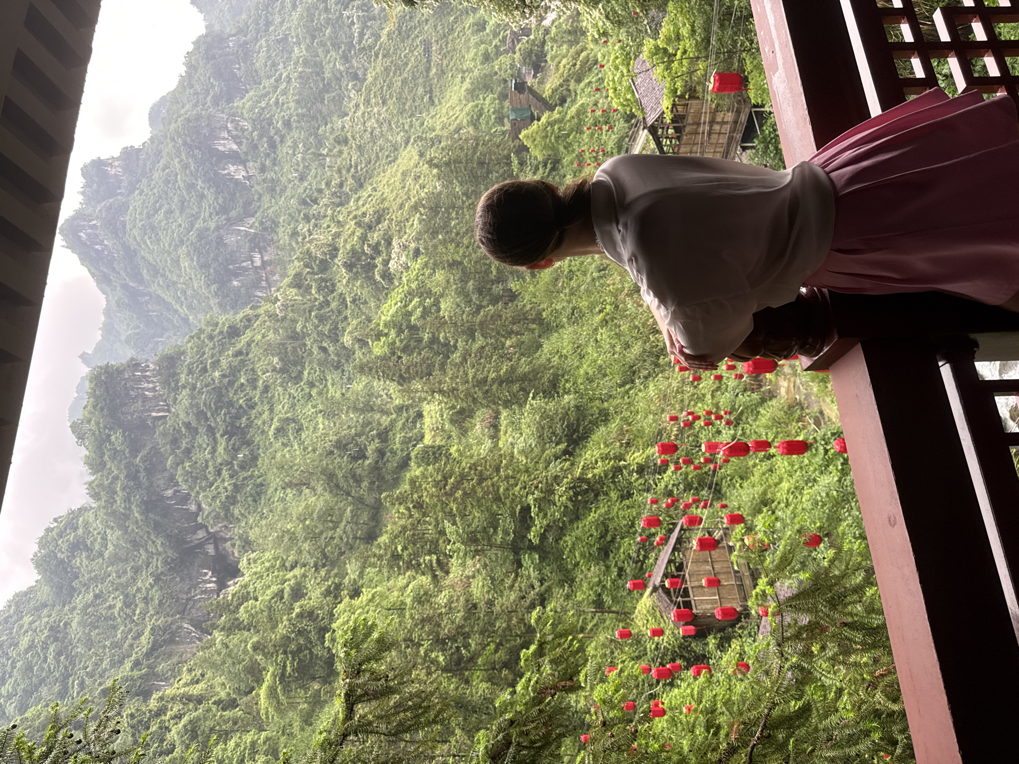 A female person with brown hair tied back is seen from behind, leaning over a wooden railing on a balcony, viewing a landscape of mountains and greenery. The person is wearing a white, long-sleeved shirt and a pink skirt. The balcony structure is made of dark wood. In the background, there are green mountains with some visible rock faces. A dense forest of trees covers the lower areas, interspersed with red lanterns. There is a small wooden structure with a peaked roof, suspended amidst the trees and lanterns. The location is most likely in Yongding District, China. The sky is overcast and the light is diffused, suggesting a misty or cloudy day.