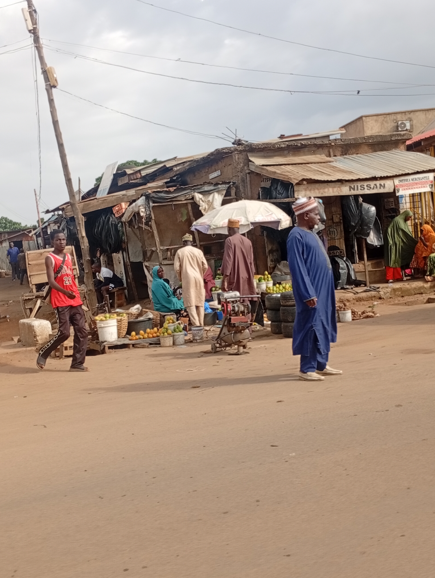 The media file depicts a busy street scene in Rafin Par, Nigeria. Several people are present, including men and women dressed in traditional attire. A young man in a red basketball jersey and black pants walks towards the foreground. In the background, several market stalls are set up along the roadside, displaying various goods. One stall appears to be selling fruits, with baskets of oranges and other produce visible. Another stall has a sign that reads "NISSAN." The structures in the background seem to be makeshift, with corrugated metal roofs and wooden supports, indicating a market or informal commercial area. A power pole with electrical wires stretches across the top of the frame. The weather appears to be overcast, with a cloudy sky. The overall atmosphere suggests an active market day.