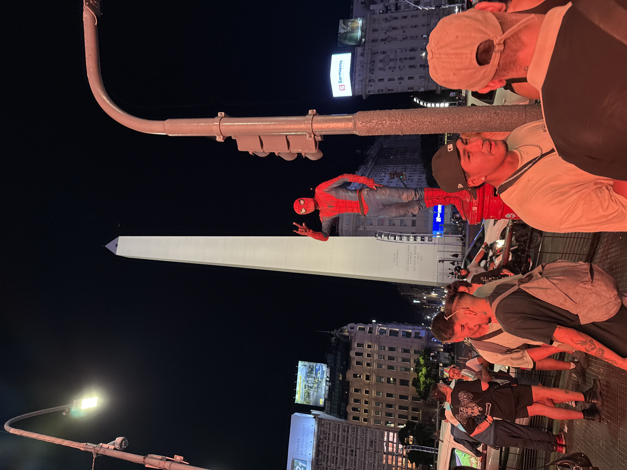 The image depicts a nighttime scene at the Obelisco in Buenos Aires, Argentina. The Obelisco, a tall white monument, is prominently featured in the background, illuminated against the night sky. In the foreground, several people are gathered, some taking pictures or observing. A person dressed as Spiderman is posing near the top of a traffic light pole, extending one arm in a friendly gesture. The surrounding environment includes city buildings illuminated with advertisements and streetlights. The general atmosphere appears lively, suggesting a public event or gathering.
