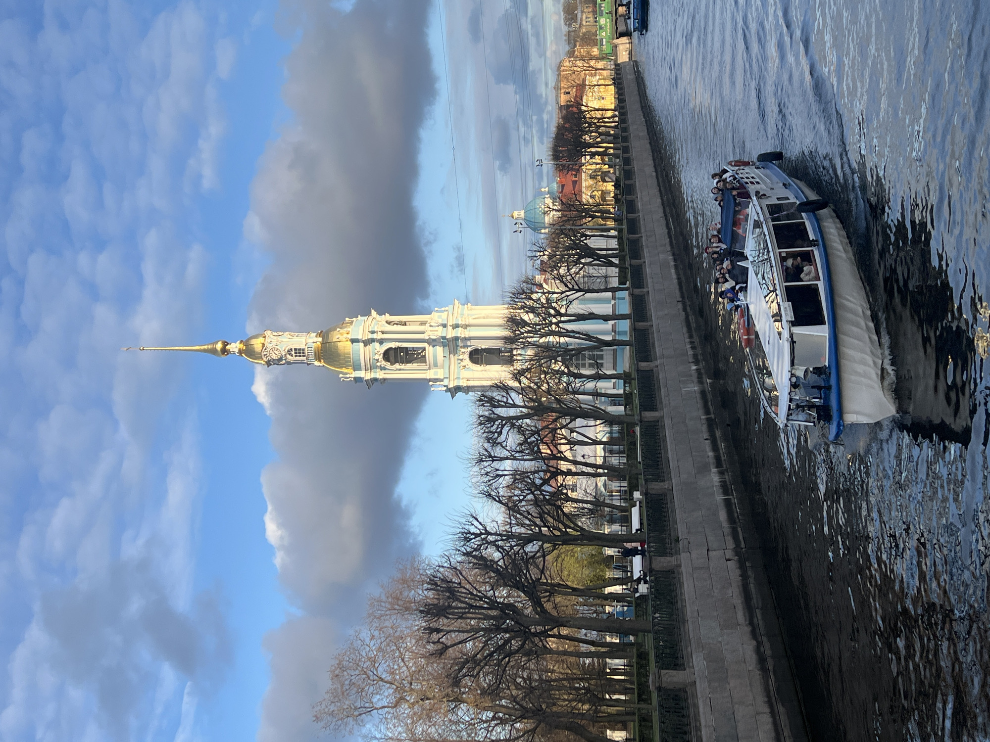 The photograph presents a view of Saint Petersburg, Russia. The Peter and Paul Cathedral, characterized by its golden spire, dominates the left side of the frame. A canal, presumably a part of the Neva River, runs alongside a stone embankment lined with bare trees and an iron fence. A tourist boat with passengers is seen on the water. The sky above is partly cloudy.