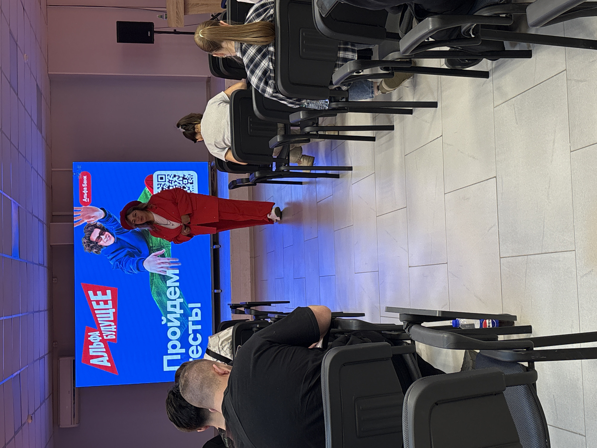 A medium shot depicts a presentation by Alfa Bank taking place indoors, possibly in a meeting or conference room in Birobidzhan, Russia. The presenter, dressed in a red pantsuit and hat, stands in front of a large screen featuring an advertisement for Alfa Bank's "Future" program. The screen is blue with red and white text and images of people. Attendees are seated in rows of black chairs, facing the screen. Some attendees are visible in the foreground and background, with varying hairstyles and clothing. The room has tiled flooring and neutral-colored walls. A bottle of water is seen on a chair's table. A speaker is visible at the top of the frame.