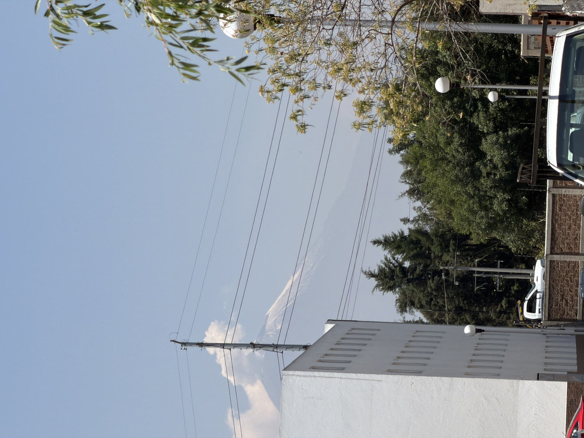 The image depicts a street scene in San Andrés Cholula, Mexico, characterized by a mix of urban and natural elements. In the foreground is a white building with evenly spaced, narrow vertical openings along its facade. Overhead, a network of power lines stretches across the sky, supported by utility poles. Behind the building and utility lines, there are dense green trees. A vehicle is partially visible on the right edge. The sky is clear and light blue. In the distant background, there appears to be a faint outline of a mountain. The lighting suggests it is daytime.