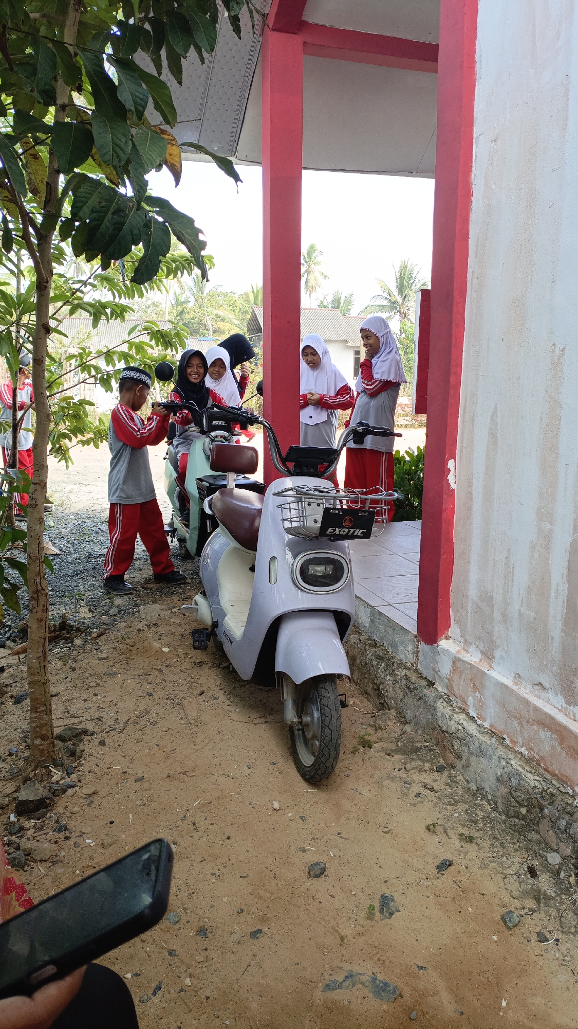 In the city of Pulau Tanjung, Indonesia, a group of students, likely in uniform, are gathered around two electric scooters. A young boy wearing a grey and red uniform and a patterned cap is interacting with one of the scooters. Behind him, three girls in white headscarves and red and grey uniforms are smiling and looking towards the camera. Another girl in similar attire stands to the right. The scooters, one teal and one light purple, are parked on a dirt and gravel ground in front of a building with red-painted pillars and a white wall. A tree with large green leaves is visible on the left. The environment appears to be an outdoor area, possibly near a school or community center, with palm trees and other vegetation visible in the background under a bright sky. A mobile phone is partially visible in the foreground.