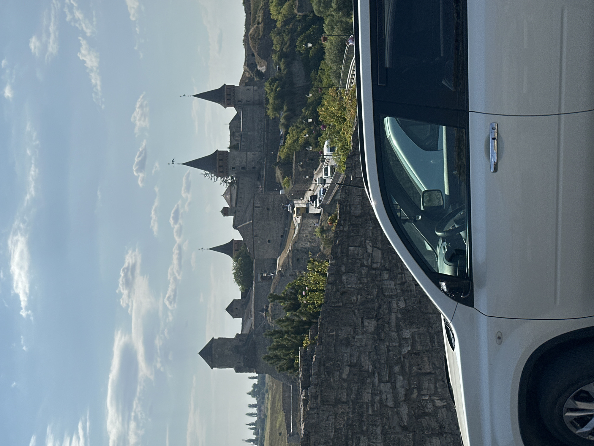 This image depicts a view of the Kamianets-Podilskyi Castle in Ukraine, taken from the perspective of a white vehicle parked in the foreground. The castle, a historic fortress, is situated on a hill overlooking a bridge where traffic is visible. The castle itself features multiple towers with pointed roofs, characteristic of its medieval architecture. Lush greenery surrounds the castle and its ramparts, with trees and bushes dotting the landscape. The sky above is a bright blue with scattered white clouds, indicating a clear day. The foreground is dominated by the side of a white SUV, with its door handle and window visible. The stone wall of a fortification runs along the lower left portion of the image, separating the vehicle from the path leading to the castle.