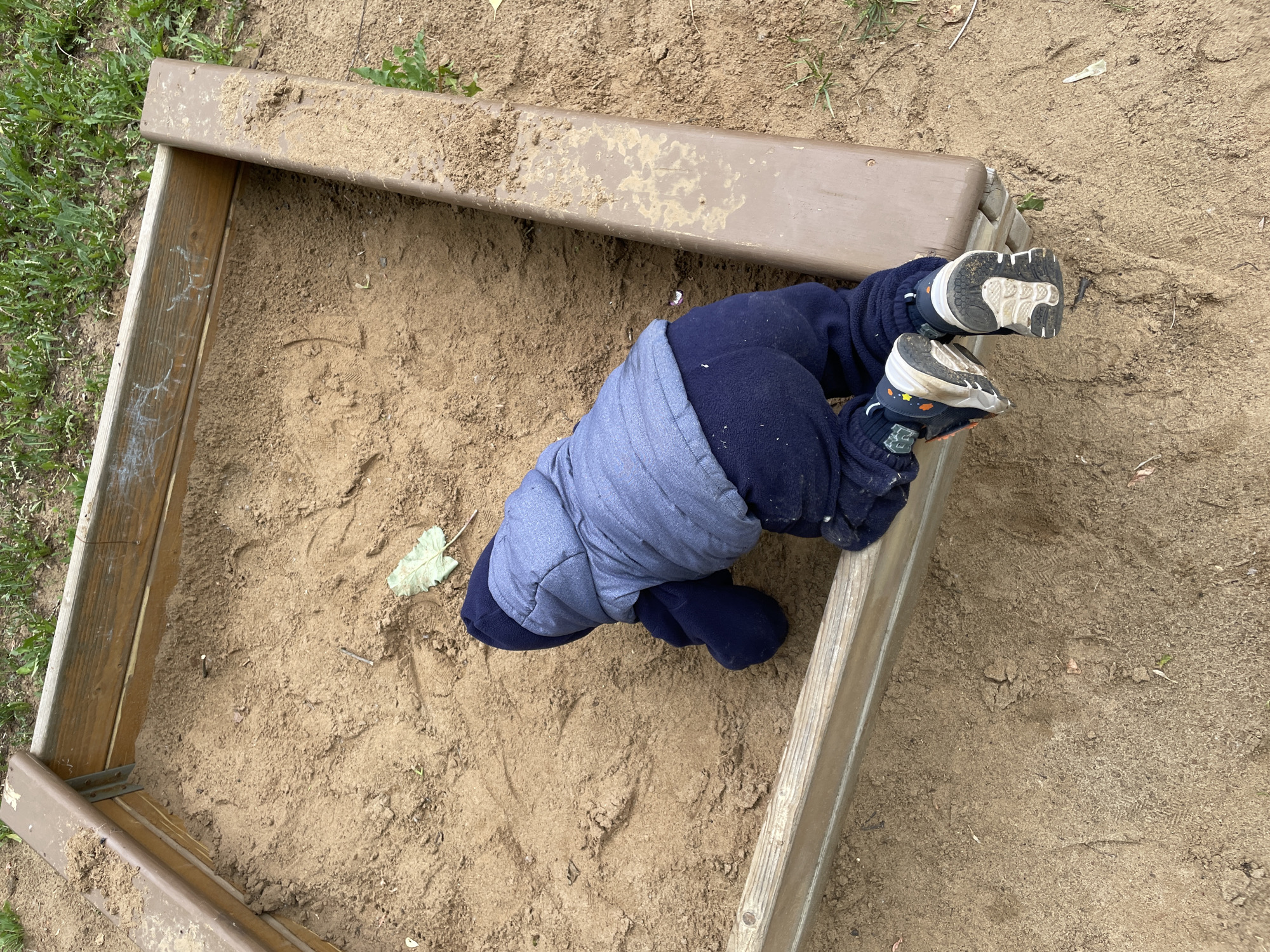 An overhead view captures a toddler, dressed in a blue hooded jacket, dark blue pants, and dark sneakers, in the act of entering or crawling within a sandbox. The child is positioned on hands and knees, facing away from the camera, with the head obscured by the hood. One leg is draped over the frame of the sandbox, while the other is fully inside. The sandbox, located in the city of Perm, Russia, features a frame constructed with a mix of light brown wooden planks and a darker brown, possibly composite, border. The interior is filled with light brown sand, which shows various indentations and contains small natural debris, including a pale green leaf and a thin stick. The area immediately surrounding the sandbox consists of sparse green grass and bare soil.