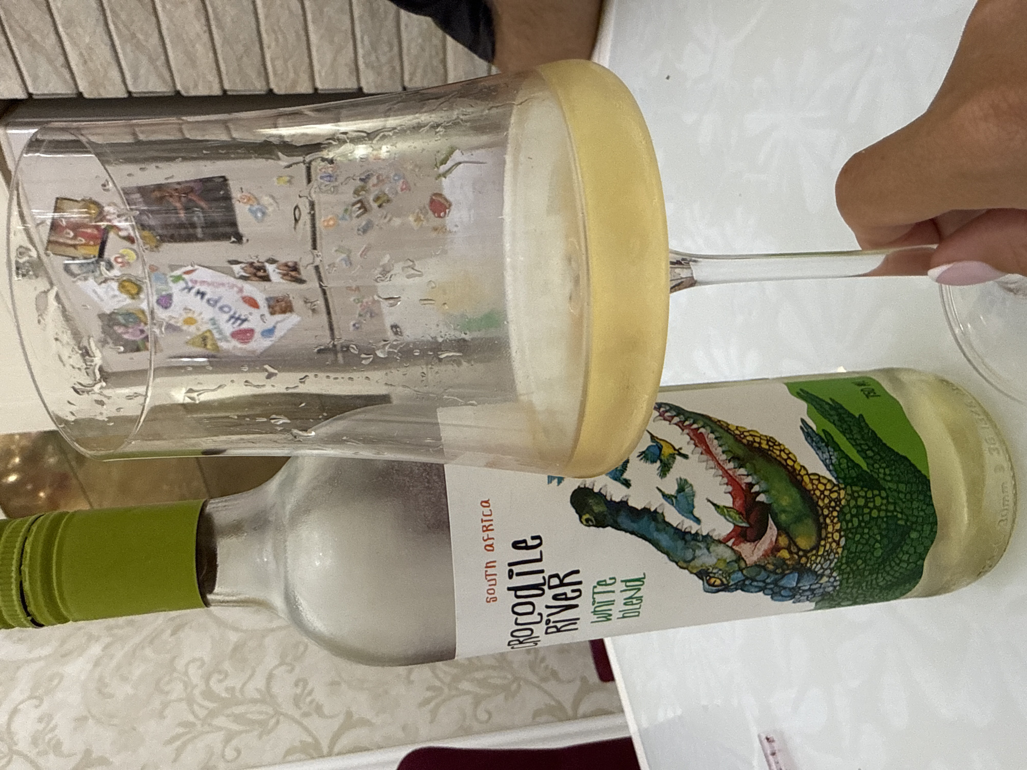 The image shows a bottle of "Crocodile River" white blend wine from South Africa and a partially filled wine glass. The bottle is lying on its side, with the label clearly visible. The glass is positioned above the bottle, suggesting that wine was recently poured. The wine glass is partially covered in condensation. A hand is partially visible holding the stem of the wine glass. The background is a patterned surface, possibly a table cloth or textured wall. There's no information in the photo to infer that the location is Sochi, Russia.