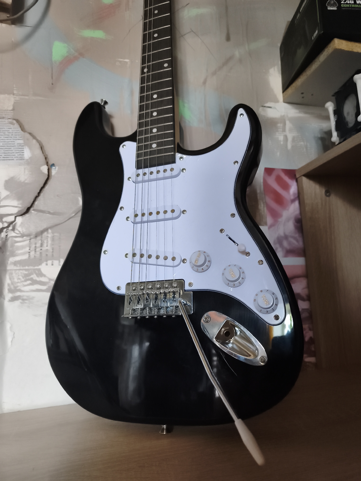 A black electric guitar with a white pickguard is positioned vertically against a wall with graffiti and torn paper. The guitar is on a wooden surface, possibly a shelf or table, with a wooden cabinet or shelf unit to the right. The guitar has six strings, a silver bridge and tailpiece, and three white control knobs labeled "VOLUME" and "TONE". A tremolo arm is inserted into the tailpiece. In the background, on a shelf above and to the right of the guitar, is a black box with a green logo. The scene appears to be indoors, possibly a room or studio.