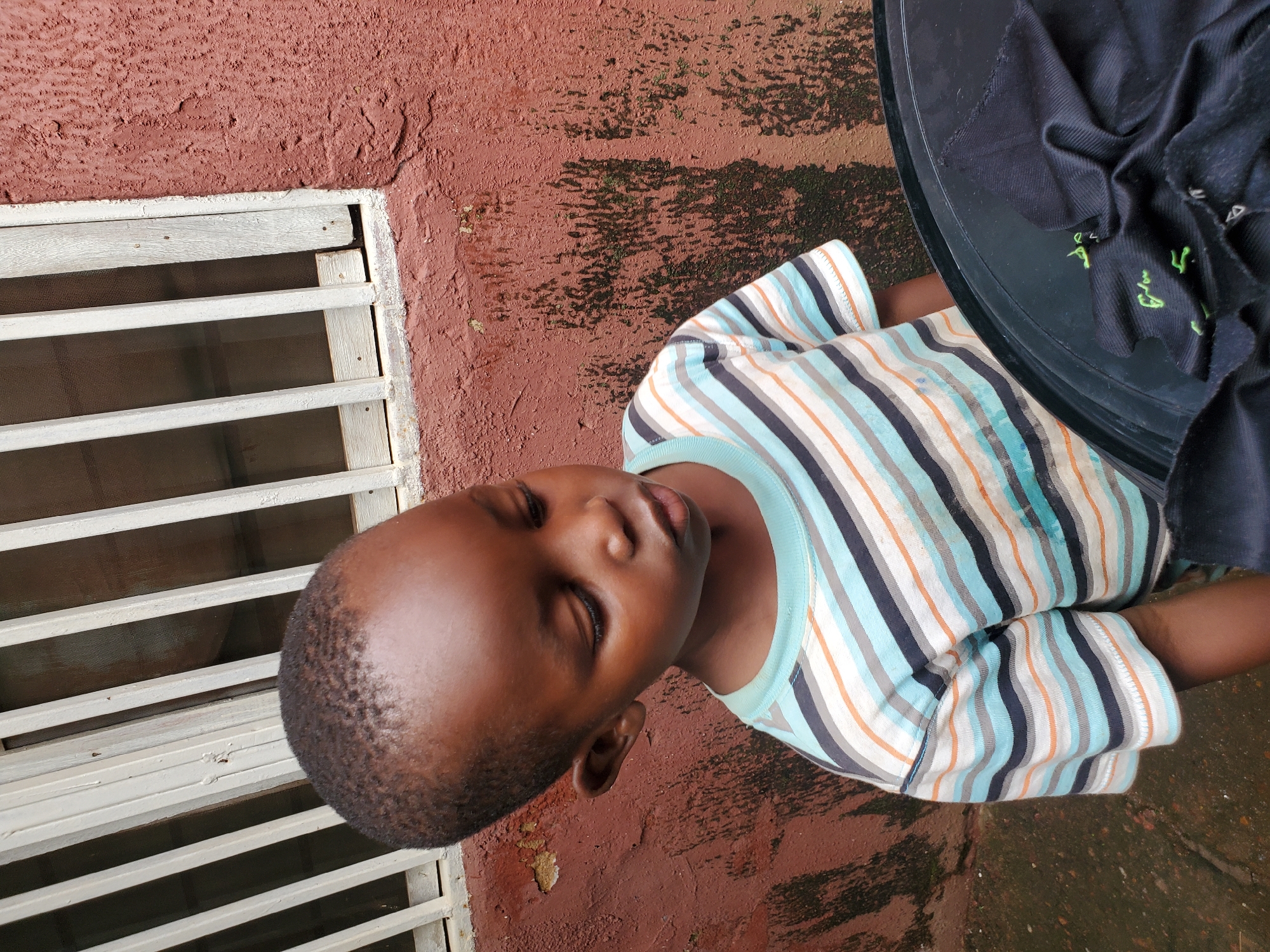 A young Black boy stands outdoors, looking down and to his right. He is wearing a horizontally striped t-shirt with alternating bands of white, light blue, dark blue, gray, and orange. The t-shirt has a light blue collar. Behind him, a reddish-brown textured wall is visible, with some green algae growth in patches. To the boy's left, a window with white bars and white trim is present. The ground appears to be wet and dirty. In the foreground, a black plastic object, possibly a drum or container, is partially visible, with some black fabric draped over it. The scene is set in Jos, Nigeria.