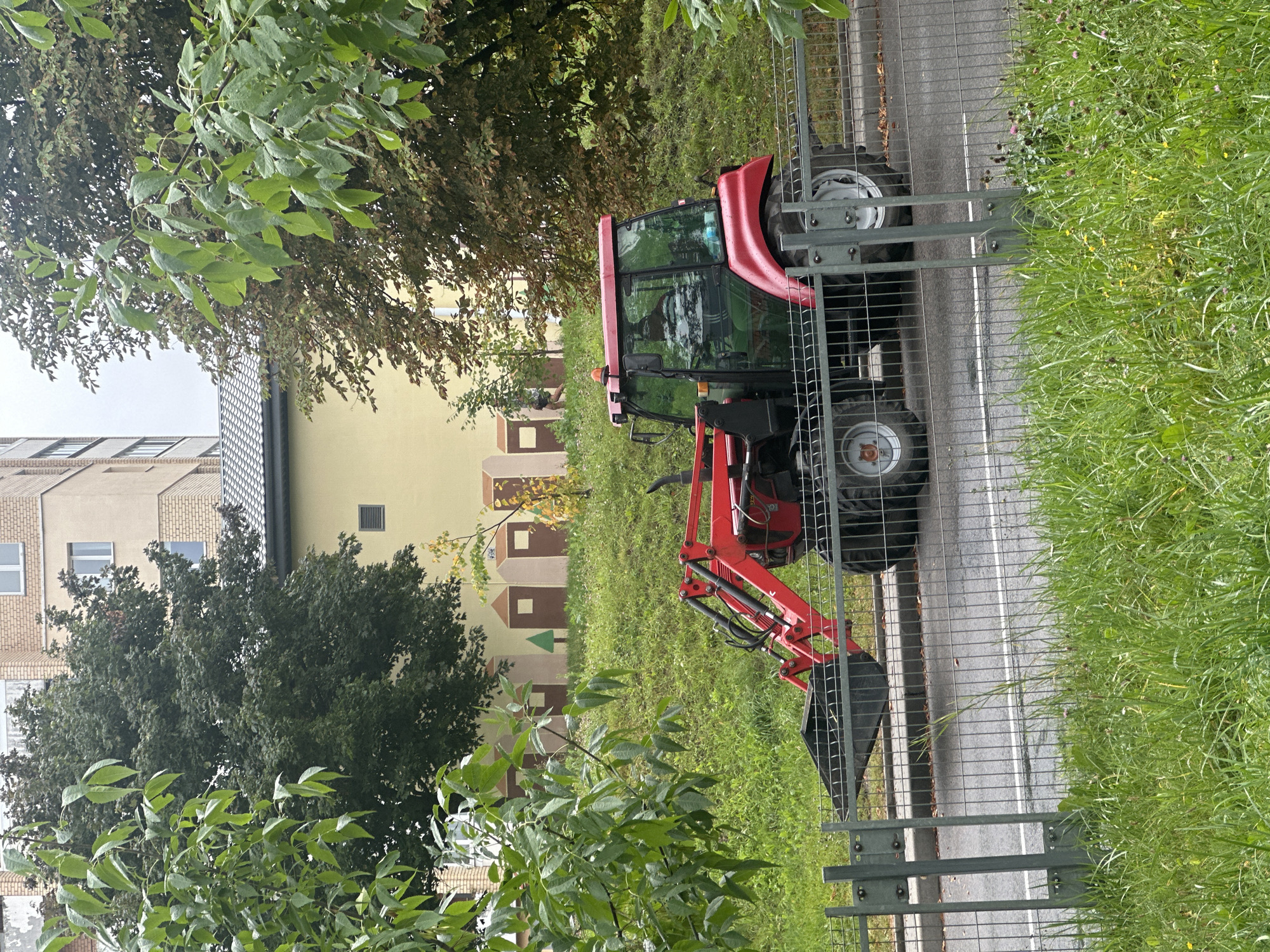 The image shows a red tractor with a front loader parked on a grassy embankment next to a street. The tractor is viewed from the side, and its tires and bucket are visible. Behind the tractor, there is a building with a yellow facade featuring a mural of stylized houses. Further behind, a multi-story apartment building is visible. Trees with green and autumn foliage are present in the foreground and background. A metal fence separates the embankment from the street. The overall scene appears to be in an urban or suburban environment, likely in Minsk, Belarus, given the context provided. No people are clearly identifiable in the scene.
