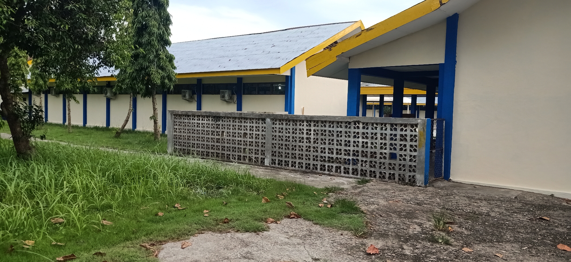 The image shows a building with a light cream-colored exterior, accented with blue trim on columns and window frames, and a yellow overhang along the roofline. The roof is made of corrugated metal. In front of the building, there is a decorative concrete block fence with a repeating geometric pattern. To the left of the fence is a patch of green grass with scattered fallen leaves. Several trees with lush green foliage are also visible to the left, with their trunks partially obscured by the grass. In the background, beyond the fence and building, another structure with a similar architectural style is partially visible. The sky is overcast. The scene appears to be an outdoor area of a facility in Tambontunong, Indonesia.