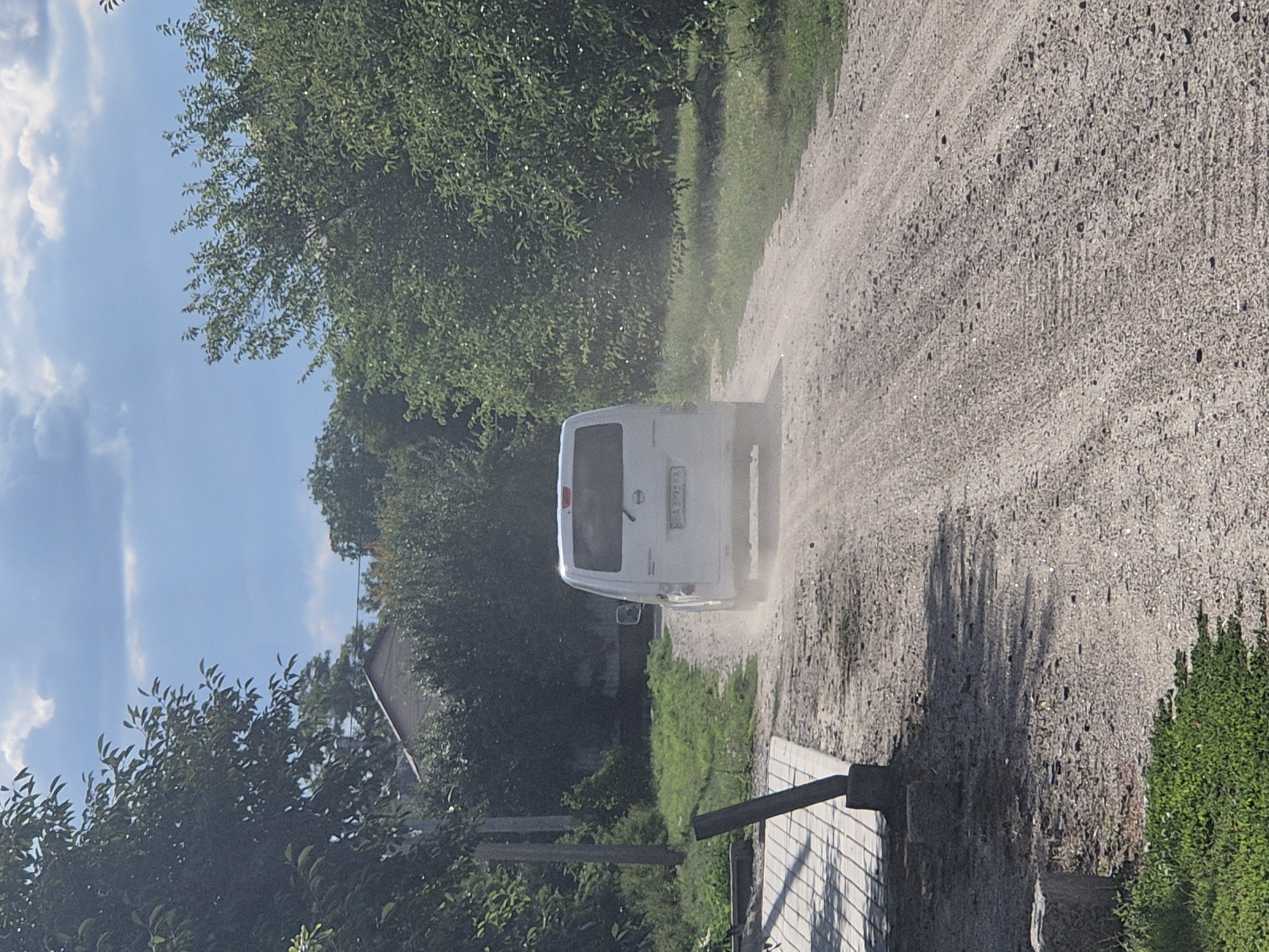 A white van is seen driving on a dirt road, kicking up dust. The van, with a license plate that appears to read "EA 3077 EC", is moving away from the viewer. The scene is set in the village of Sozonivka, Ukraine, as indicated by the visible environment. Lush green trees and foliage line the sides of the road, with a house visible in the background through the trees. A paved area with cobblestones and a dark pole are visible on the left side of the road, suggesting a pathway or sidewalk. The sky is a mix of blue with scattered clouds. The overall impression is a rural road on a sunny day.
