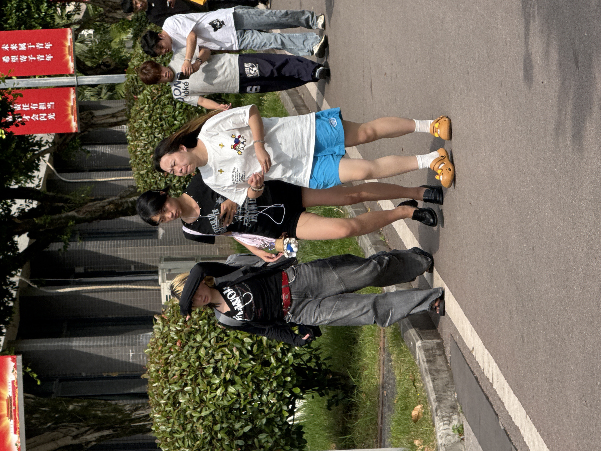 The image shows a group of people walking along a road near a green hedge in what appears to be an urban environment. Red signs with Chinese writing are visible in the background; one sign states, "Hope is placed on the youth. The future belongs to the youth." This suggests the location is in China, specifically Jiangjin.
There are approximately six individuals in the scene. Some are wearing casual attire like t-shirts and jeans. One person wears a white t-shirt with blue shorts and yellow shoes. Another person is wearing black clothing with gray jeans and a red belt, appearing to carry flowers. They seem to be walking or moving together.
The background also features buildings and trees, further indicating a cityscape setting. The photograph is taken during the daytime with clear sunlight.