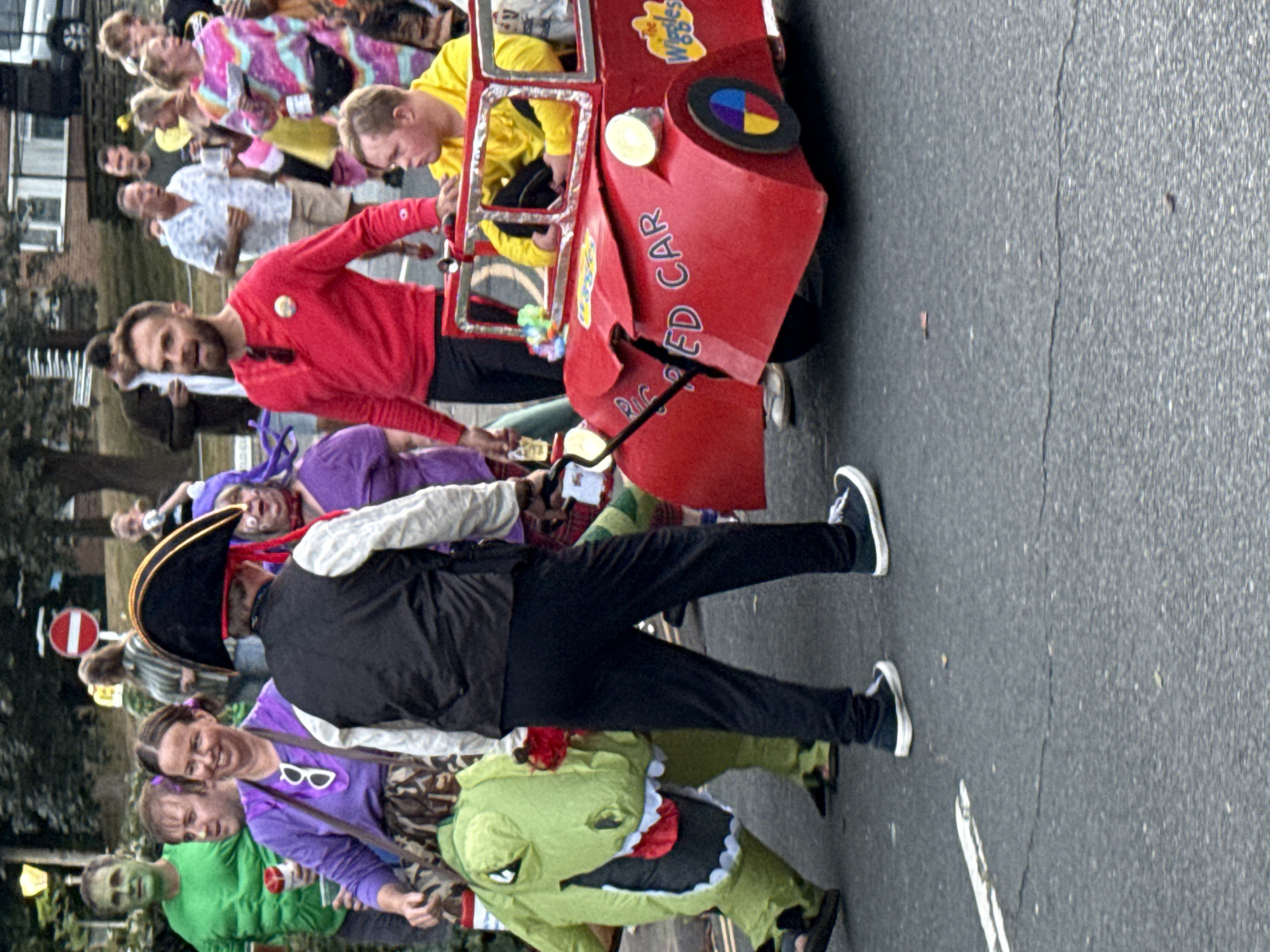 In St Leonards, United Kingdom, on a street, a group of people in costumes are participating in an event, possibly a parade. In the foreground, a person in a pirate costume stands next to a child in a yellow shirt inside a homemade red toy car. The car is decorated with "Big Red Car" and "The Wiggles" logos. To the left, a person in a green dinosaur costume walks. Other individuals in various colorful outfits are visible in the background, including one in a purple outfit with a blue hairpiece. The street is paved and has a white line marking. Trees and buildings are present in the background.