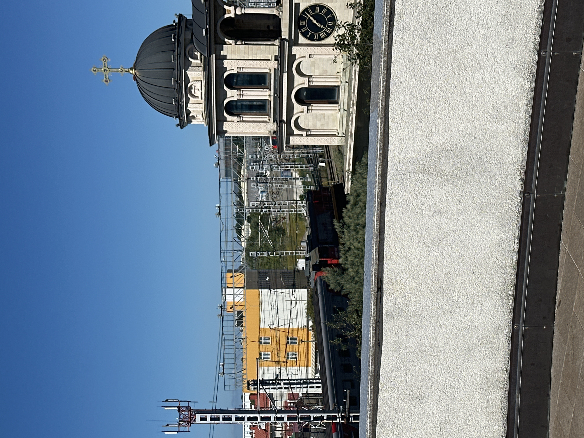 This is a photograph of the city of Sirius, Russia, captured on a clear day with a bright blue sky. In the foreground, a textured white wall is visible, with a darker border at the bottom. Beyond this wall, the scene opens up to reveal several structures and elements.
To the right, a prominent building with a dark dome topped by a golden cross stands out. The building is constructed of light-colored stone and features arched windows and decorative elements. A clock face with Roman numerals is visible on one side of the building.
In the mid-ground, a train with a red front is seen on railway tracks. Adjacent to the tracks, there are several buildings. One building is prominently yellow and white, with multiple windows. To the left of this building, a tall, red and white cell tower is situated. Various wires and scaffolding are also present in this area.
Further in the background, more buildings and structures can be observed, along with greenery and a hilly landscape. The overall impression is an urban environment with a mix of religious, industrial, and residential elements.