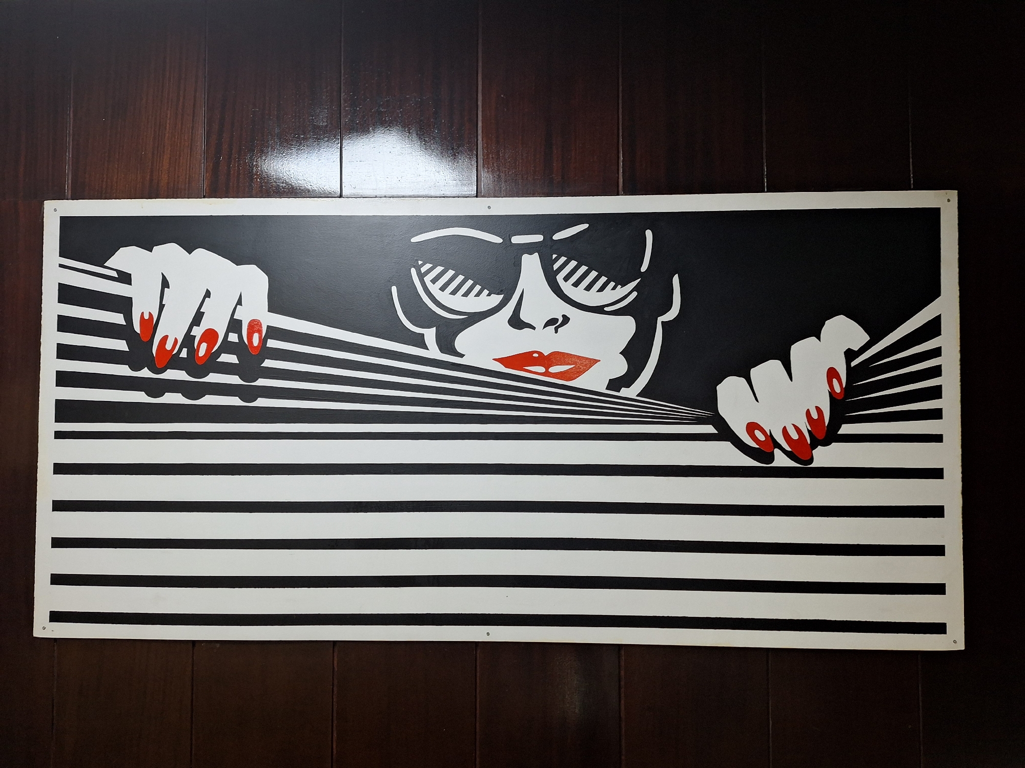 The image is a close-up shot of a framed painting hanging on a dark brown wooden wall. The painting is rectangular and predominantly white with black details. The artwork depicts a stylized face with dark sunglasses, red lips, and hands pulling apart black and white horizontal stripes, creating the illusion of blinds being opened. The fingernails are painted bright red. The wall is made of vertical wooden panels with a glossy finish, reflecting light near the top. The painting is attached to the wall with metal pins in the corners. The location in the city of Varna, Bulgaria is not detectable from the image.