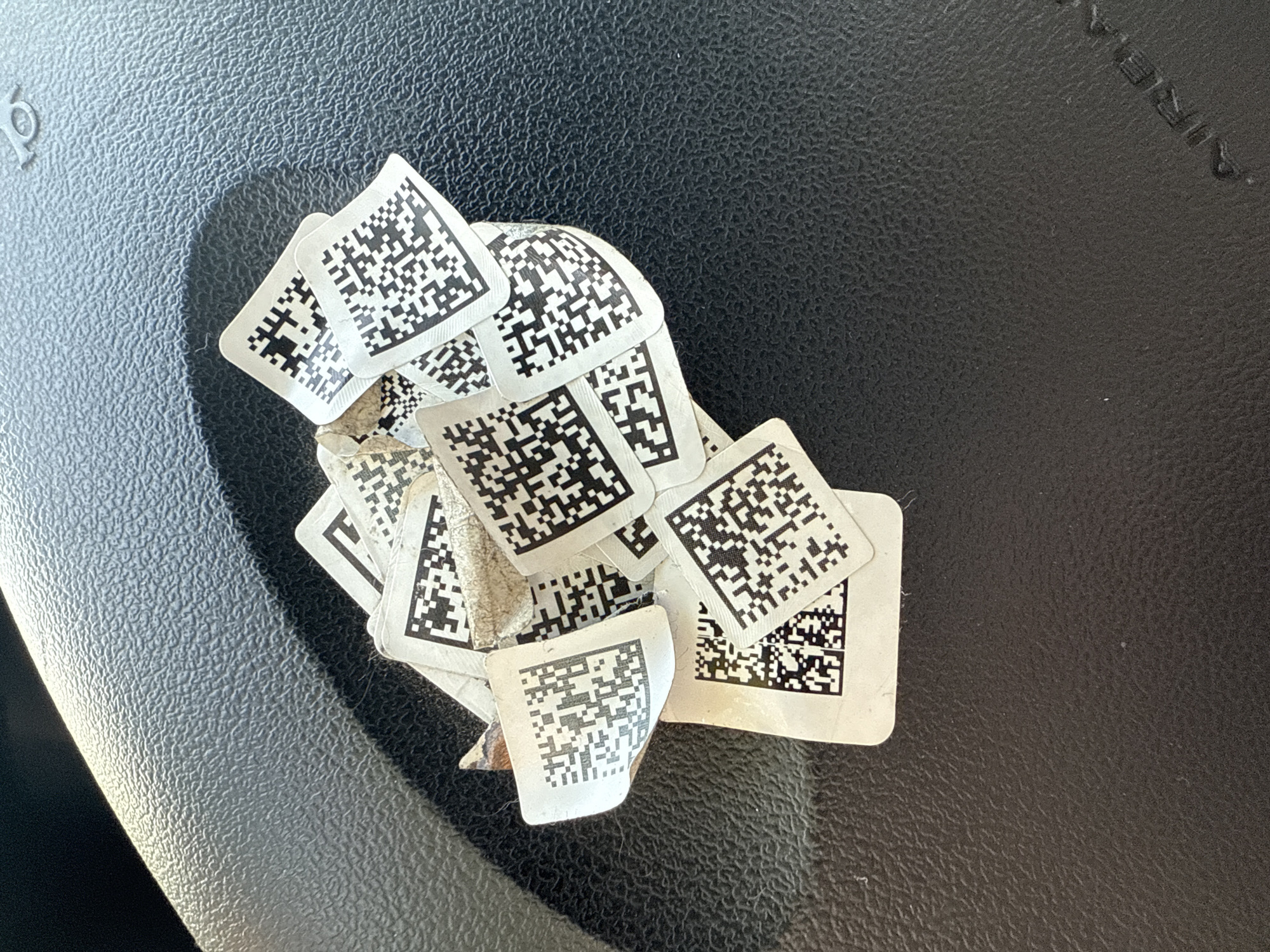 A close-up, overhead shot shows a pile of small, white stickers with black QR codes on them. The stickers are scattered and layered on top of each other, with some appearing torn or creased. The background is a dark, textured surface, likely a steering wheel, with a curved, lighter grey object in the upper left corner. The lighting suggests an indoor or shaded outdoor environment, with a distinct shadow cast across the surface. There is no indication of the specific location in Samara, Russia, or any people or actions within the frame.