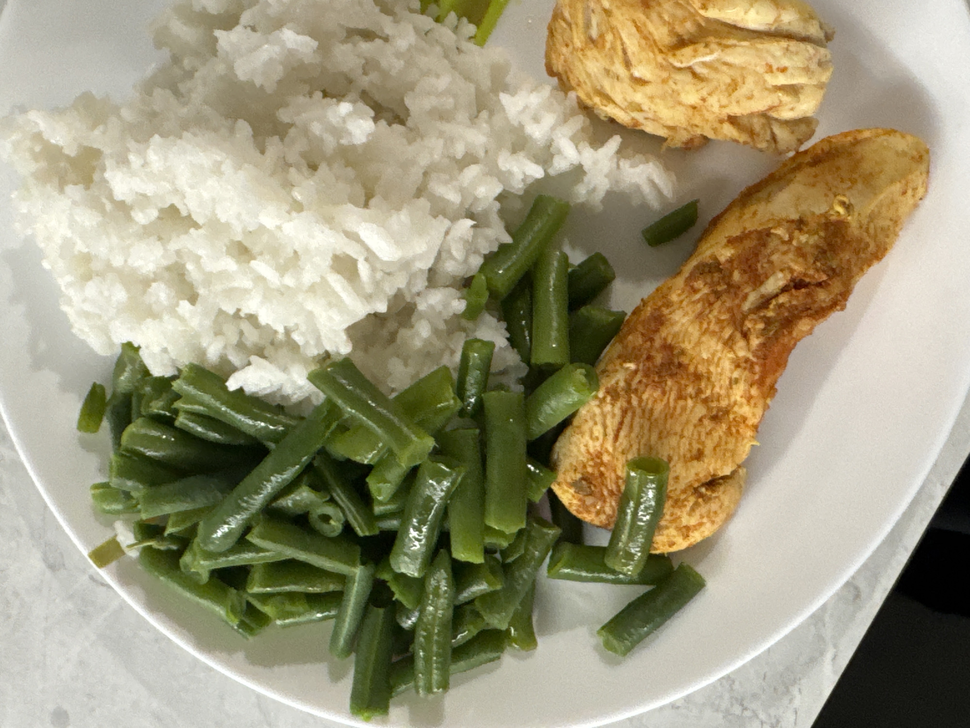 A white plate containing a meal is seen on a light gray surface. The meal consists of a mound of white rice, a pile of green beans, and two pieces of cooked chicken. The chicken pieces are golden brown. The image does not contain enough information to determine the specific location in Chaikovsky, Russia. There are no people visible in the image.