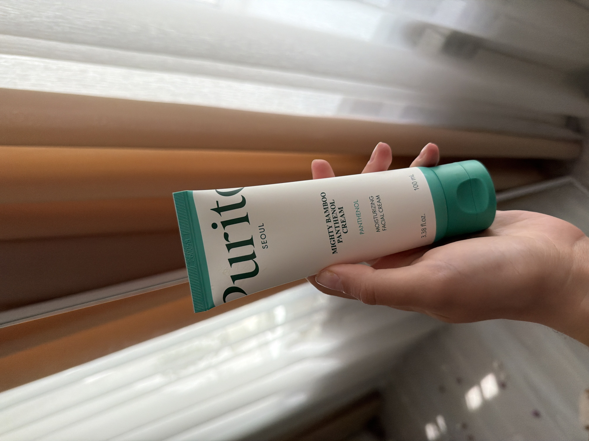 A hand holds a white tube of Purito "Mighty Bamboo Panthenol Cream" with green lettering and a teal cap. The tube is positioned vertically, with the cap at the bottom. The background consists of sheer white curtains and a brown curtain. The lighting suggests an indoor setting, possibly near a window. The image does not contain any information that allows for determining its location in Semiluki, Russia.