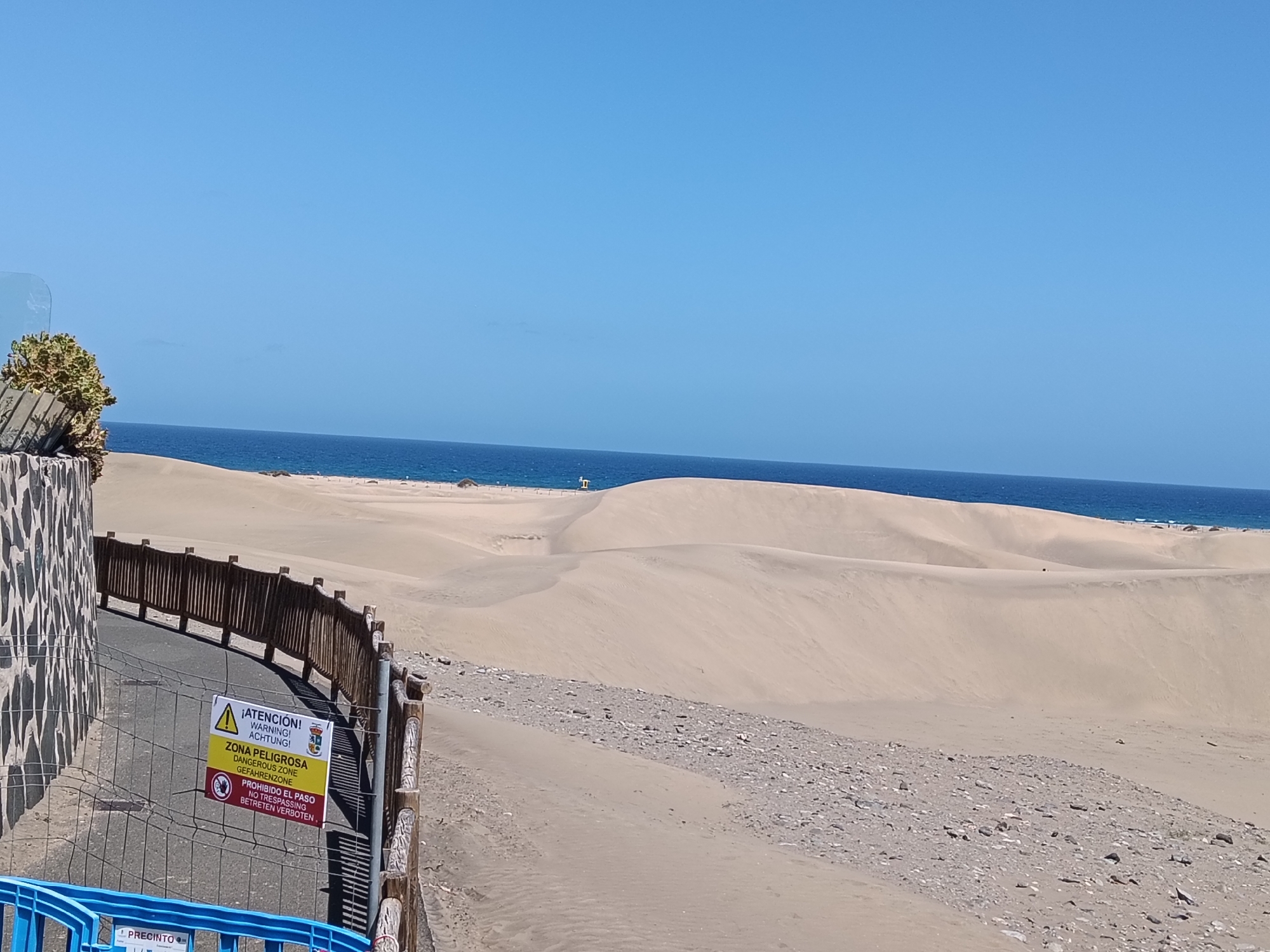 The image depicts a sunny, outdoor scene featuring the Maspalomas Dunes in San Bartolomé de Tirajana, Spain. The foreground includes a partially visible stone wall and a wooden fence. A sign attached to the fence reads "¡ATENCIÓN! WARNING! ACHTUNG!" with the warning "ZONA PELIGROSA DANGEROUS ZONE GEFAHRENZONE" and "PROHIBIDO EL PASO NO TRESPASSING BETRETEN VERBOTEN." A blue plastic barrier is also visible.
The midground is dominated by the sand dunes, with varying textures and colors from light to dark beige. In the background, a stretch of the blue ocean meets the clear blue sky.