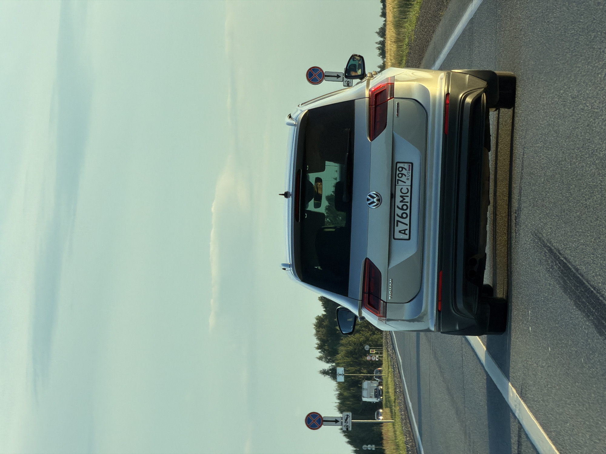 The image shows the rear view of a silver Volkswagen Tiguan driving on a highway. The car has a license plate with the registration number A766MC 799, indicating it is from Russia. In the background, there are trees and a field of wheat, suggesting a rural or semi-rural setting. There are also traffic signs visible on the left side of the road, including a "No Stopping" sign and a sign indicating a tow-away zone. The sky is partly cloudy, and the lighting suggests it is either early morning or late afternoon. The image appears to be taken from another vehicle on the same highway, possibly following the Tiguan. The location is identified as Obukhovo, Russia.
