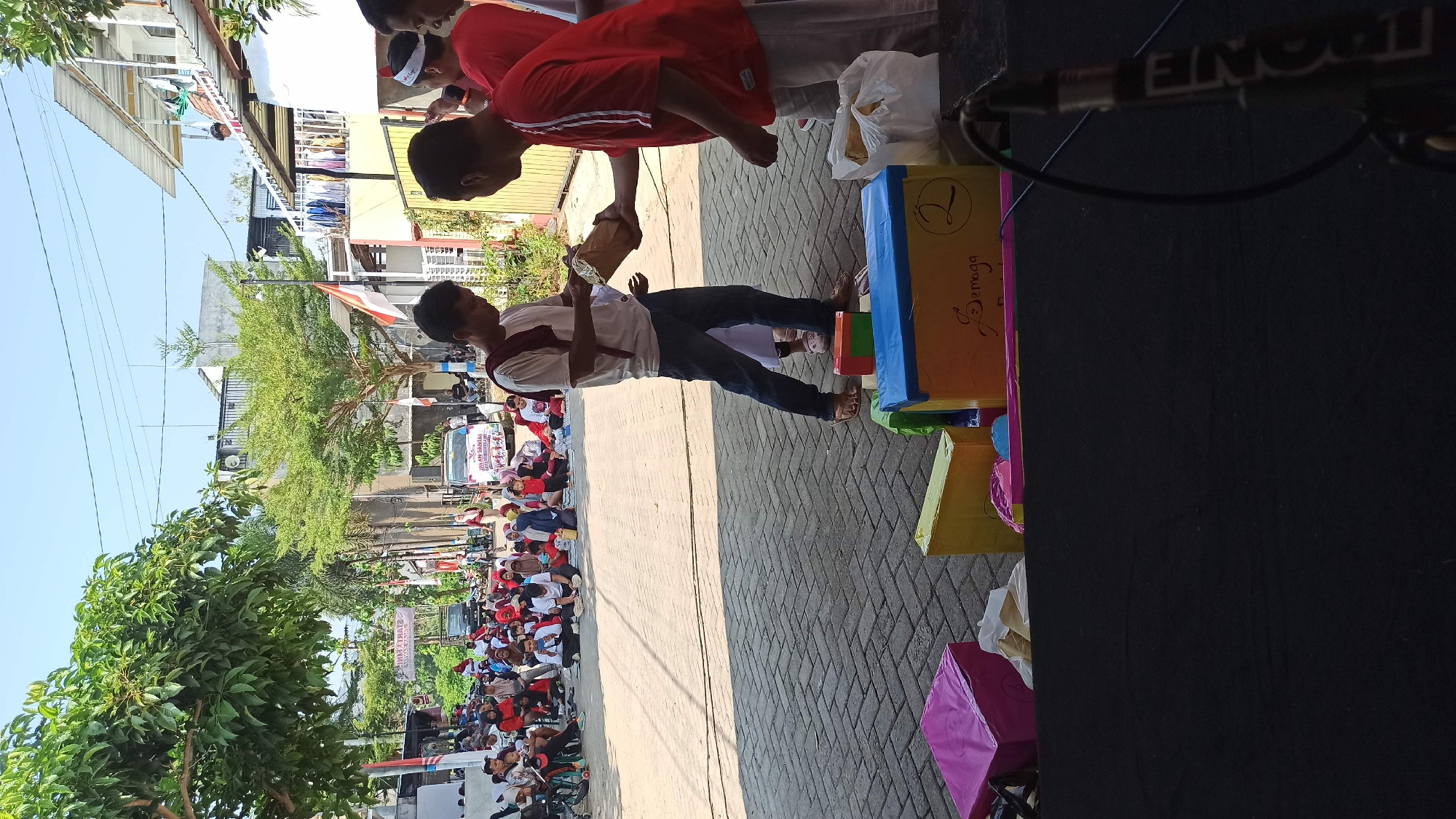 In Galak, Indonesia, a street event is underway, marked by a gathering of people sitting on the ground, watching a presentation. Two men are engaged in an exchange, one handing a wrapped object to the other. Colorful boxes are arranged on the ground, possibly prizes or props for the event. The environment suggests a sunny day, with buildings and trees framing the scene.