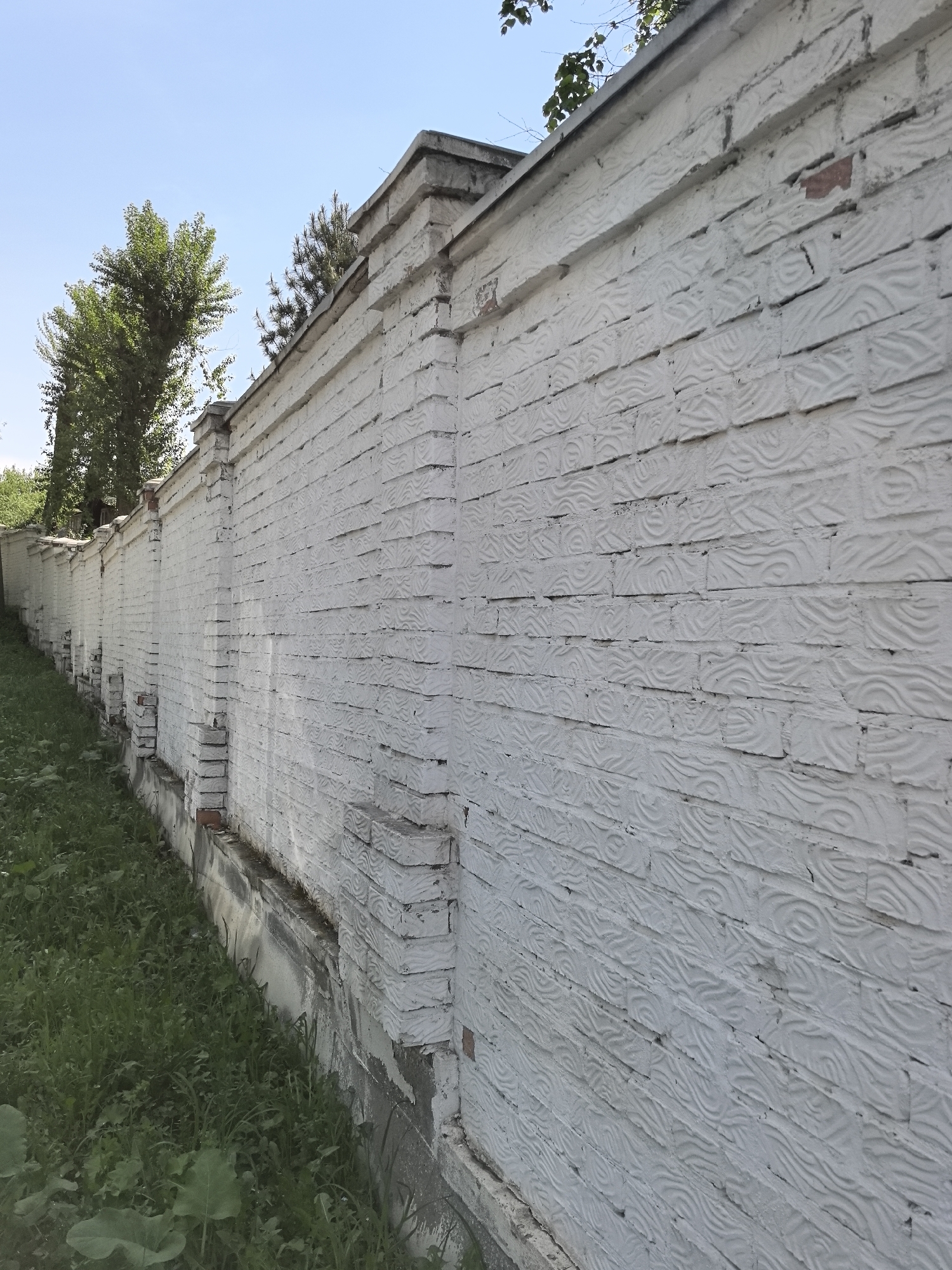 A long, white brick wall runs along a grassy area under a clear blue sky. The wall has decorative patterns on the bricks. Parts of trees are visible above the wall, and some foliage grows along the base. The location is Perm, Russia.