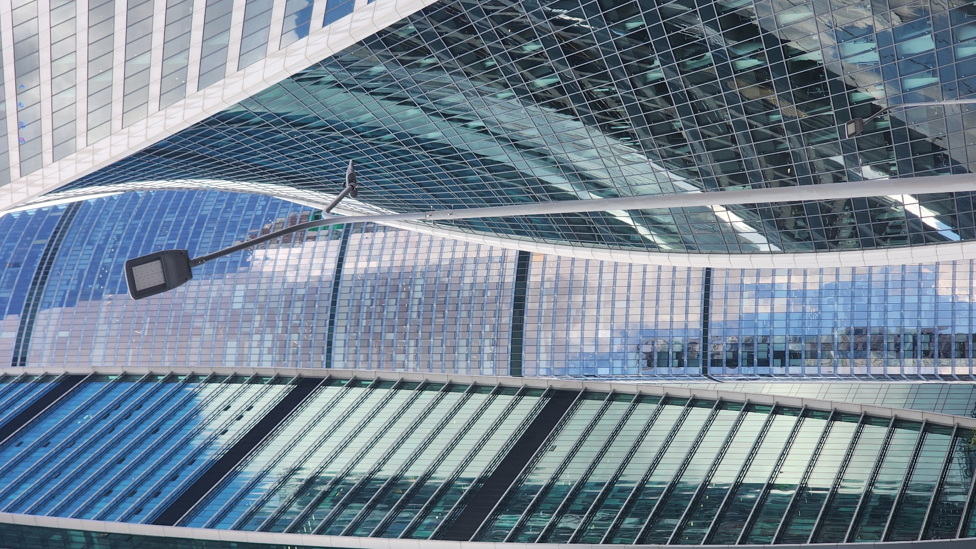 The image is a low-angle, full-frame shot of the Moscow International Business Center (MIBC), also known as Moscow-City. The scene features several modern skyscrapers with glass facades that reflect the sky and surrounding buildings. A street lamp with a modern, rectangular light fixture is prominently positioned in the foreground, slightly off-center to the right, with its pole extending vertically through the frame. The buildings exhibit contemporary architectural designs, with curved and angled surfaces, and are densely packed together, creating a sense of urban density. The lighting suggests it is daytime, with the sky appearing overcast, casting a diffused light on the structures. No people or vehicles are visible in the image. The overall impression is one of a modern, dynamic urban landscape.