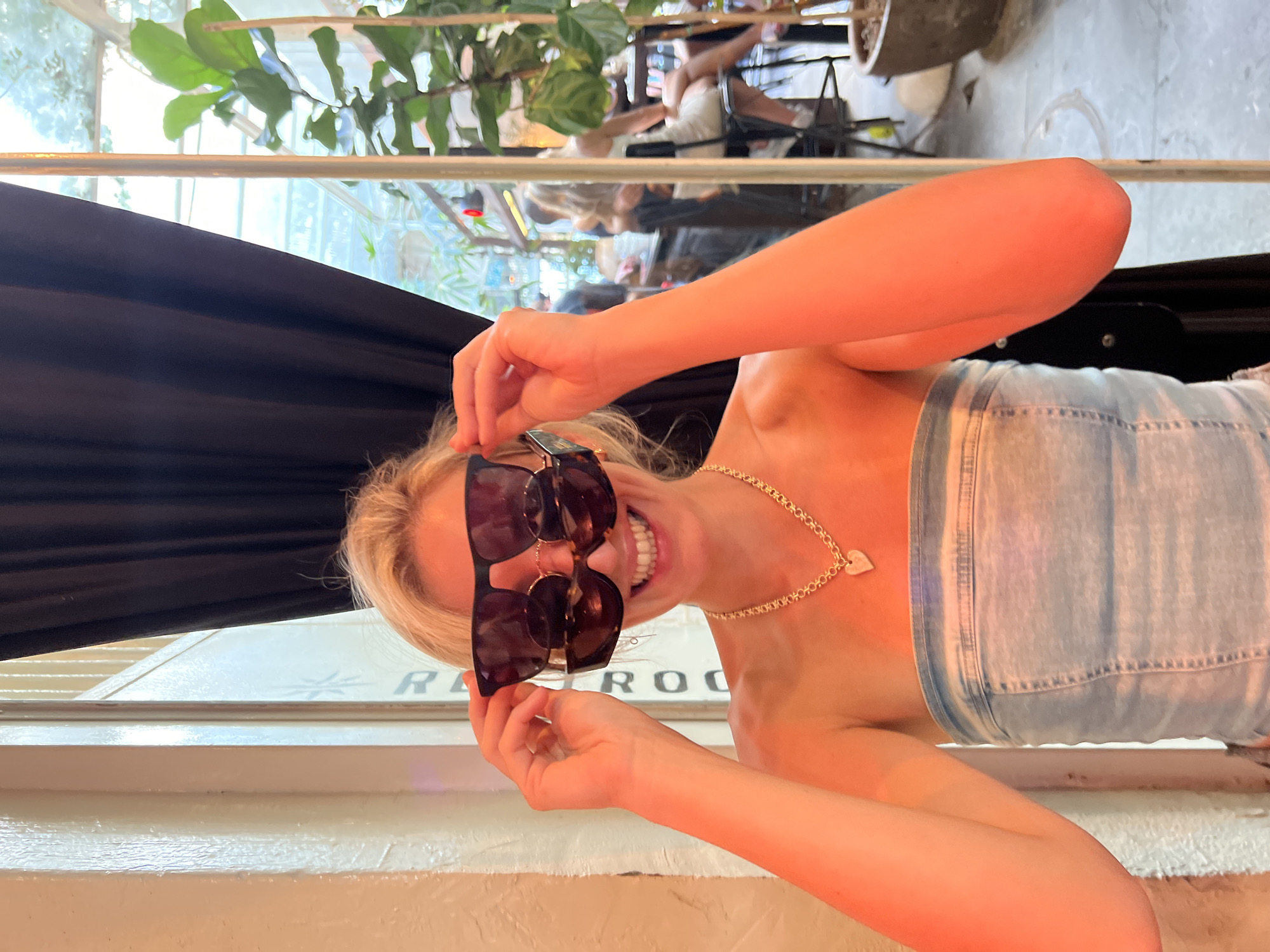 A blonde woman stands in front of a window and a black curtain, holding two pairs of sunglasses over her eyes. She is wearing a strapless denim top and a gold necklace with a heart-shaped pendant. Her arms are raised, and she is smiling. In the background, through the window, several people are visible in a dimly lit room. A large potted plant with broad leaves is located to the right of the frame. The lighting is warm and casts an orange hue on the woman. The scene appears to be in Tel Aviv, Israel, given the presence of what looks like the inscription "Room" on a sign behind the woman and the general architectural style visible through the window suggesting a venue in the city.