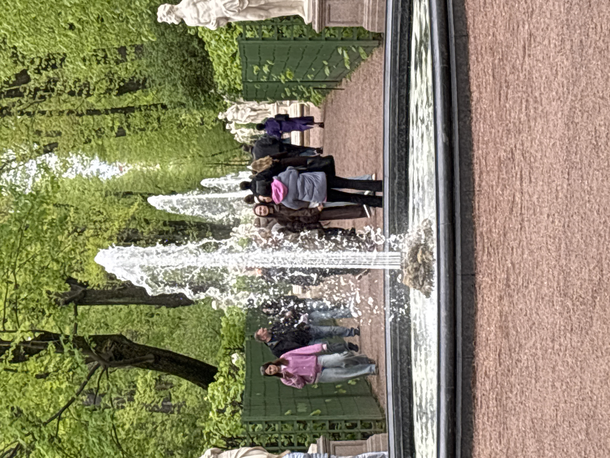 The image shows a fountain in a park, likely in Saint Petersburg, Russia. The fountain is long and rectangular, with water jets shooting upwards. People are walking along a path near the fountain. In the background, there are trees and statues. The image appears to be a reflection in the fountain's water. The right side of the image has a rough, textured surface, possibly a wall or pavement.