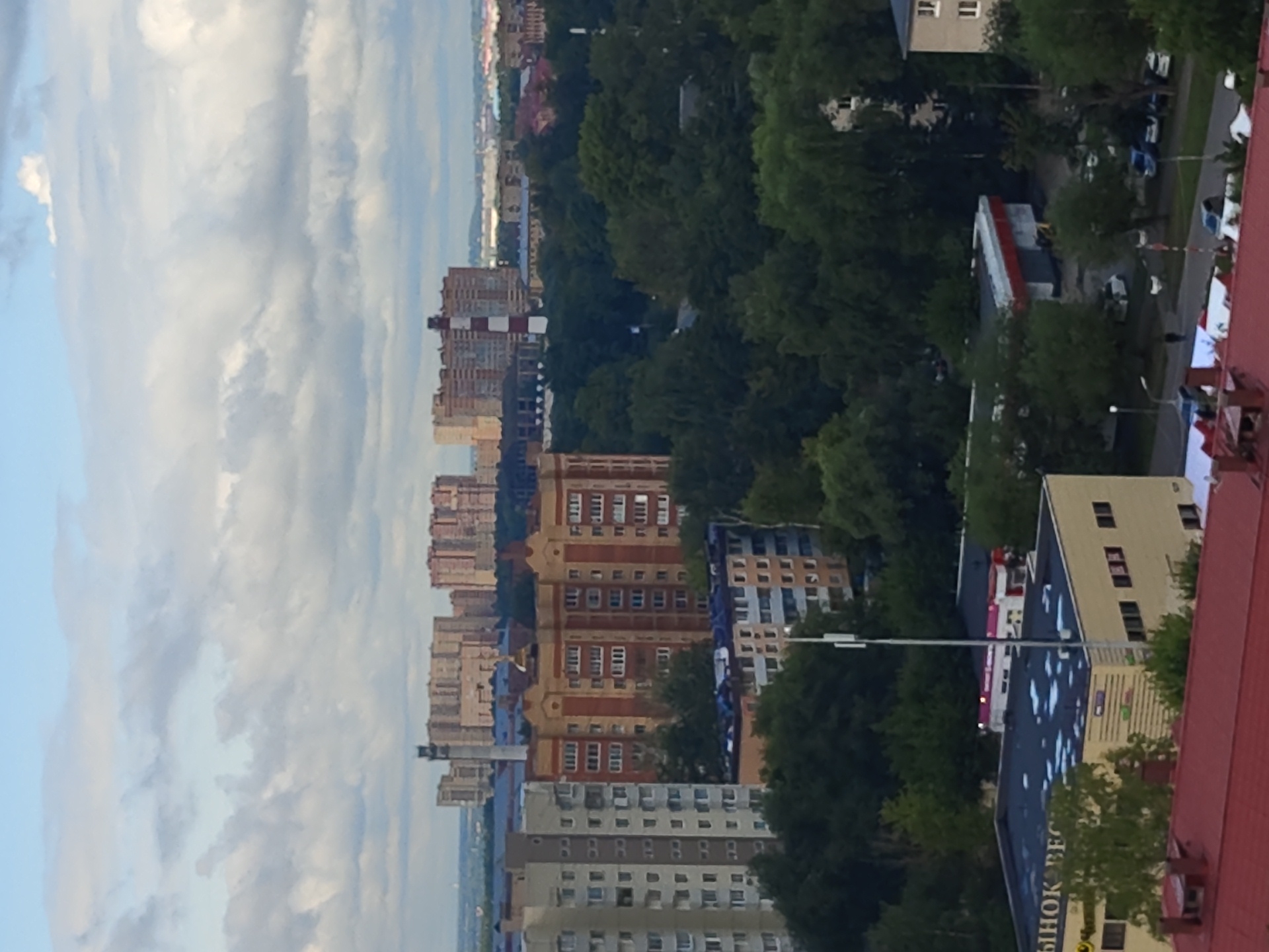The image shows a cityscape with multiple buildings and a cloudy sky. In the foreground, a red-roofed building is visible. Further back, there is a yellow building with a dark blue roof, displaying text in Russian. A tall antenna stands near this building. Beyond that, a dense canopy of green trees obscures many of the structures. Several apartment buildings of varying heights and architectural styles are scattered throughout the scene. Some buildings are light gray and tall, while others are shorter and have a brick facade with reddish-orange and yellow tones. In the distance, larger, more complex structures are visible, including what appears to be a smokestack with red and white bands. The overall impression is a residential area within a city, likely Ramenskoye, Russia, given the architectural style and the Russian text. There are no clearly discernible people or distinct actions taking place.