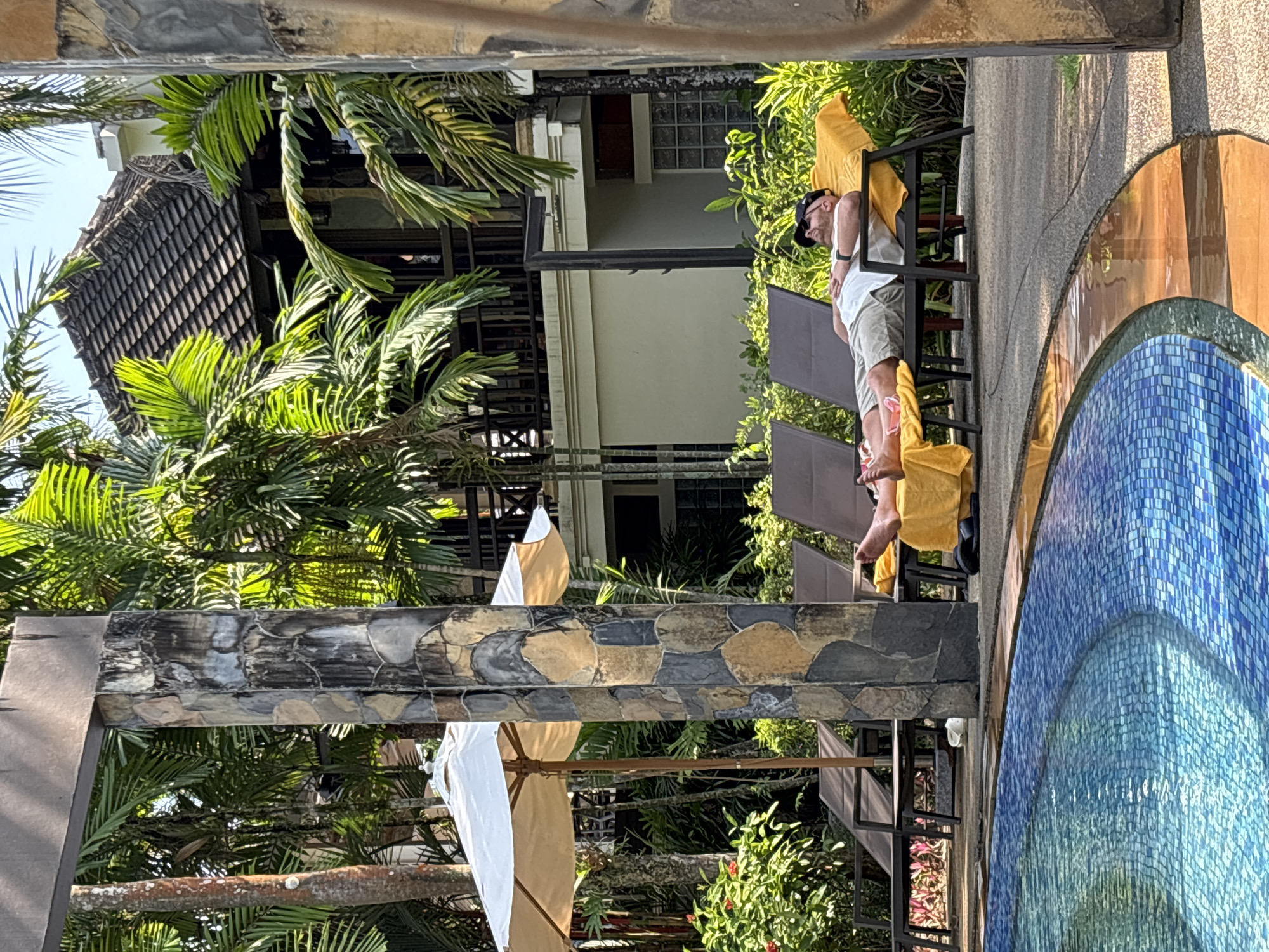 A man is sleeping on a lounge chair next to a swimming pool in Khok Kloi, Thailand. He is wearing a white shirt, khaki shorts, and a black baseball cap. The pool is tiled with blue and yellow mosaic tiles. Palm trees and a building with a brown roof are in the background. An umbrella is partially visible in the lower left corner.