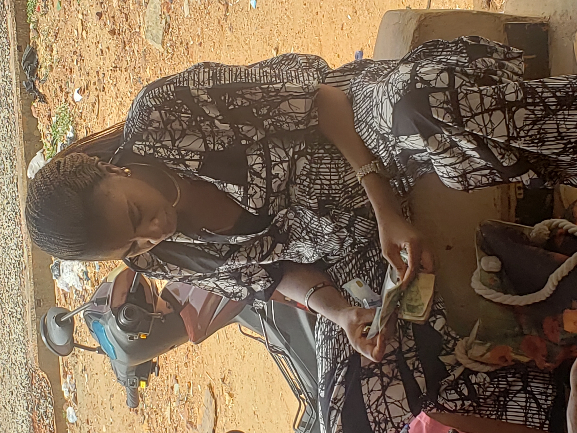 The image captures a woman handling money outdoors in Adankolo, Nigeria, during bright daylight. She is positioned with her head slightly bowed, focusing intently on the banknotes in her hands.
The woman has dark skin and her hair is styled in neat braids. She is dressed in a black and white patterned garment, possibly a traditional African print, and wears gold-colored hoop earrings and a bracelet on her left wrist. She is holding several Nigerian Naira banknotes, including green ones, which she appears to be counting, sorting, or examining.
To her left, a dark-colored scooter or motorcycle is partially visible. To her right, a dark bag or basket with a thick white rope handle rests on an unseen surface. The ground is a reddish-brown dirt, indicative of an unpaved area, and is scattered with small debris. In the background, a simple, light-colored, possibly mud-brick or concrete structure can be seen. The scene suggests a transaction or a moment of managing funds in an informal outdoor setting, likely a market, roadside, or residential area. The woman's expression conveys concentration. No specific text other than that generally found on currency is legible.
