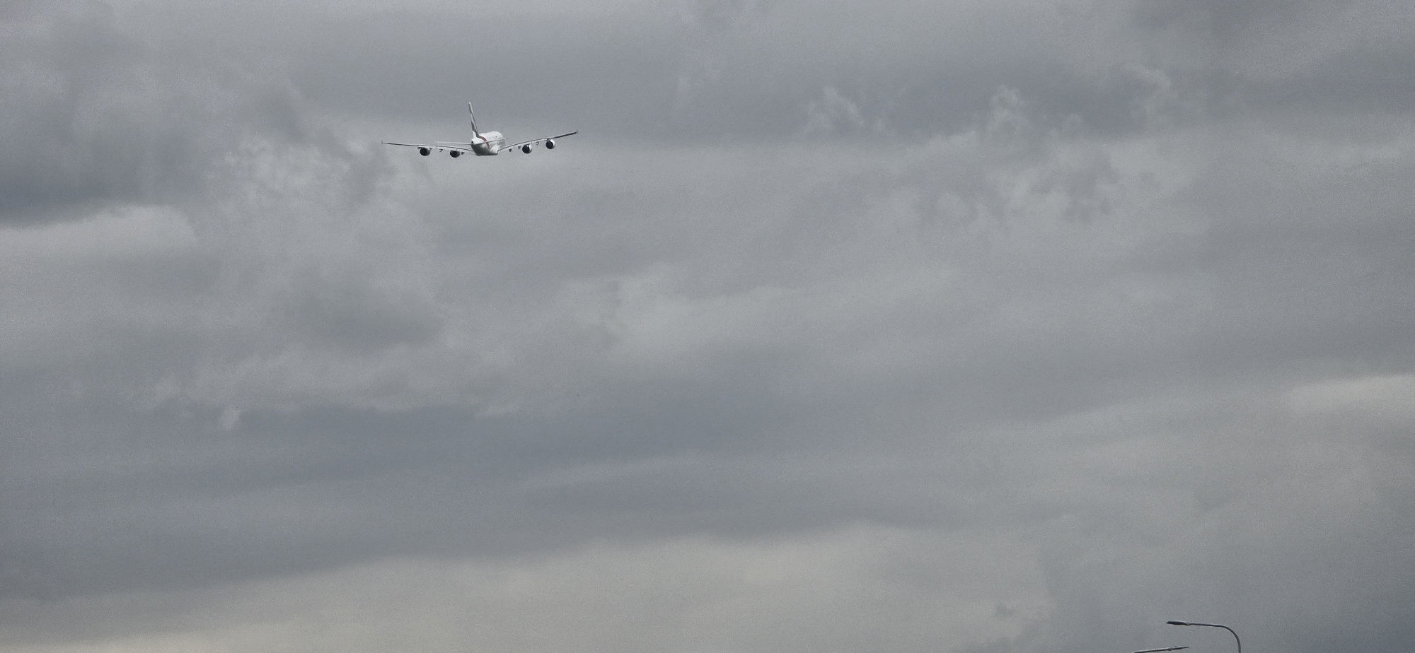 An airplane, identified as an Airbus A380 based on its distinctive four engines and double-deck configuration, is captured in mid-flight against a heavily overcast, grey sky. The aircraft is positioned in the upper left quadrant of the frame, viewed from the rear as it ascends or travels away from the viewer. The clouds are dense and uniform, with no visible breaks or patches of blue sky. The lighting conditions suggest a diffused light source, characteristic of an overcast day. In the lower right corner of the image, two streetlights with curved arms are partially visible, their presence indicating an urban or suburban environment. This scene is likely captured in the vicinity of Amsterdam Airport Schiphol (AMS), as the A380 is a wide-body airliner that operates on select routes and Schiphol is a major international airport in the Netherlands. Given the context of Hoofddorp being a town adjacent to Schiphol, this image was likely taken from Hoofddorp, Netherlands.