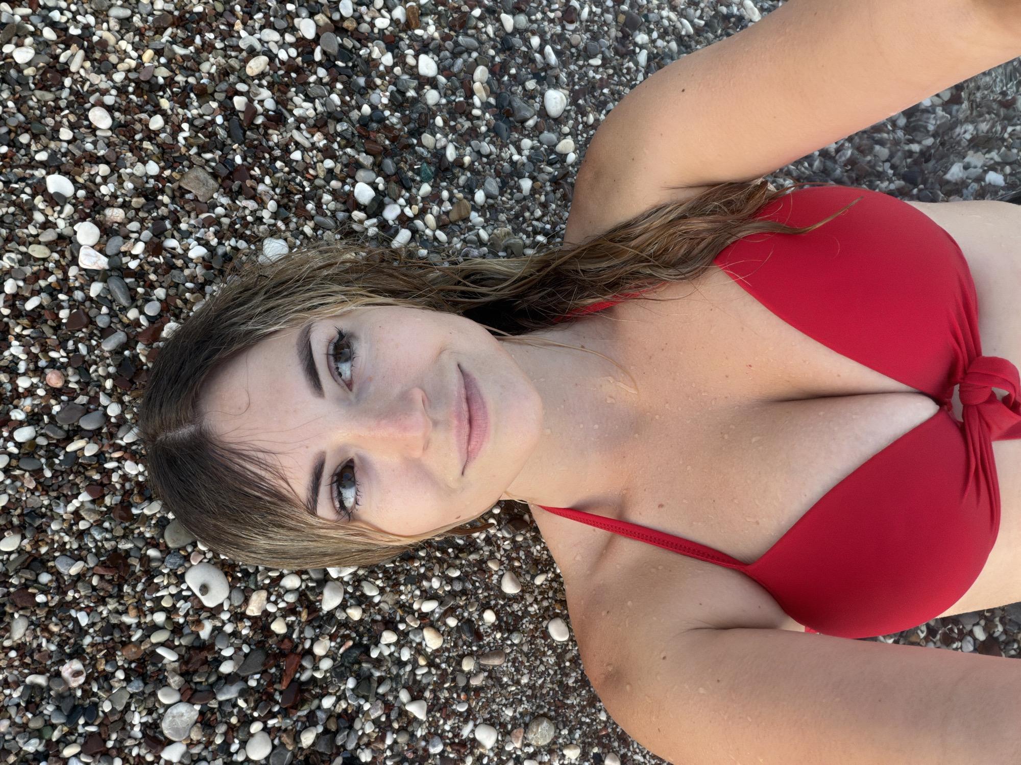 An overhead shot shows a woman lying on a pebble beach in Budva, Montenegro. She is wearing a red bikini top and has wet, light brown hair. The pebbles are a mix of gray, white, and brown colors. She is looking up and to her right with a slight smile.