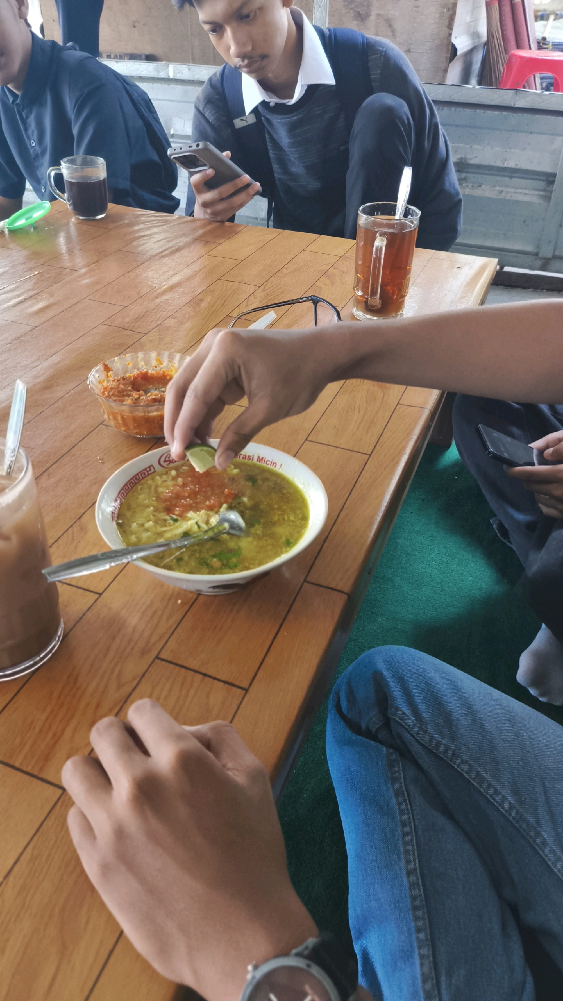 In a casual setting, likely a roadside eatery or warung in Ngemplak, Indonesia, three young men are engaged in a meal. The scene unfolds on a wooden table. In the foreground, one individual, wearing blue jeans and a watch, has their hands resting on the table. Their left hand is close to the camera, while their right hand is positioned over a bowl of soup, adding a slice of lime. The soup, served in a white bowl with red and yellow accents, contains noodles and appears to be a local delicacy. To the left of the soup bowl, a glass of iced coffee sits, partially visible. Behind the soup, a small bowl holds a vibrant red chili paste or sambal.
Further back, another young man is focused on his smartphone, which he holds in his right hand. He's dressed in a navy blue long-sleeved shirt and wears a white collared shirt underneath, suggesting a school uniform or a smart-casual attire. A backpack is slung over his shoulder. A glass of dark tea, possibly Indonesian tea, with a tea bag string hanging out, is placed to his right. The third individual, on the far left, is mostly obscured but appears to be wearing a navy blue uniform-like shirt. The background suggests an open-air or semi-open environment, with glimpses of what could be corrugated metal roofing and possibly other patrons or elements of the eatery. The overall atmosphere is relaxed and communal, typical of a casual meal shared among friends.