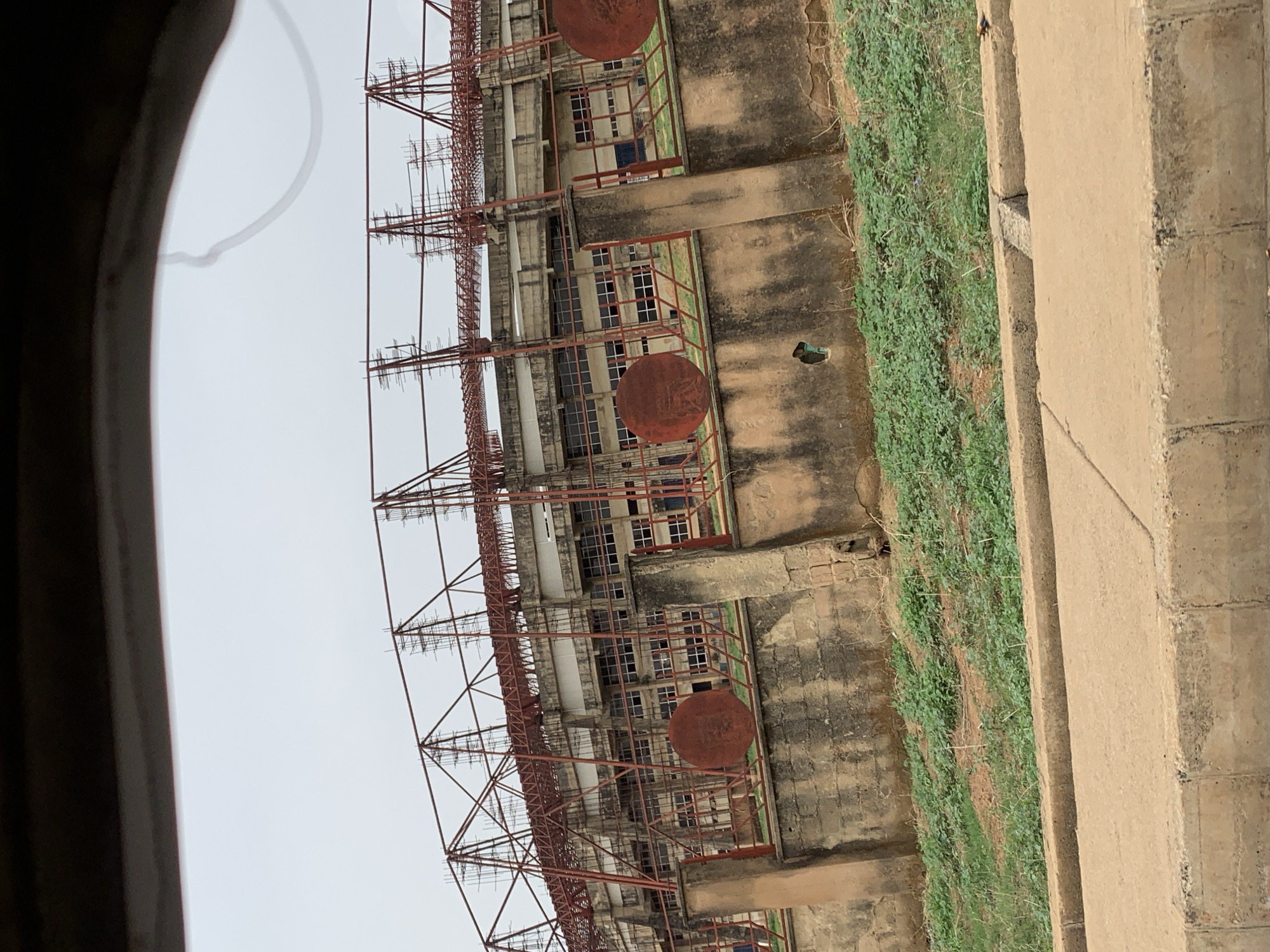 The image shows the exterior of a building in New Stadium, Nigeria. The structure appears to be an older building, possibly under construction or renovation, as evidenced by the extensive scaffolding on its side. The building has multiple windows and concrete supports along its facade. Patches of green vegetation grow along the base of the building. A concrete wall or structure is visible on the right side of the frame. The sky is overcast. The image is taken from an elevated perspective.