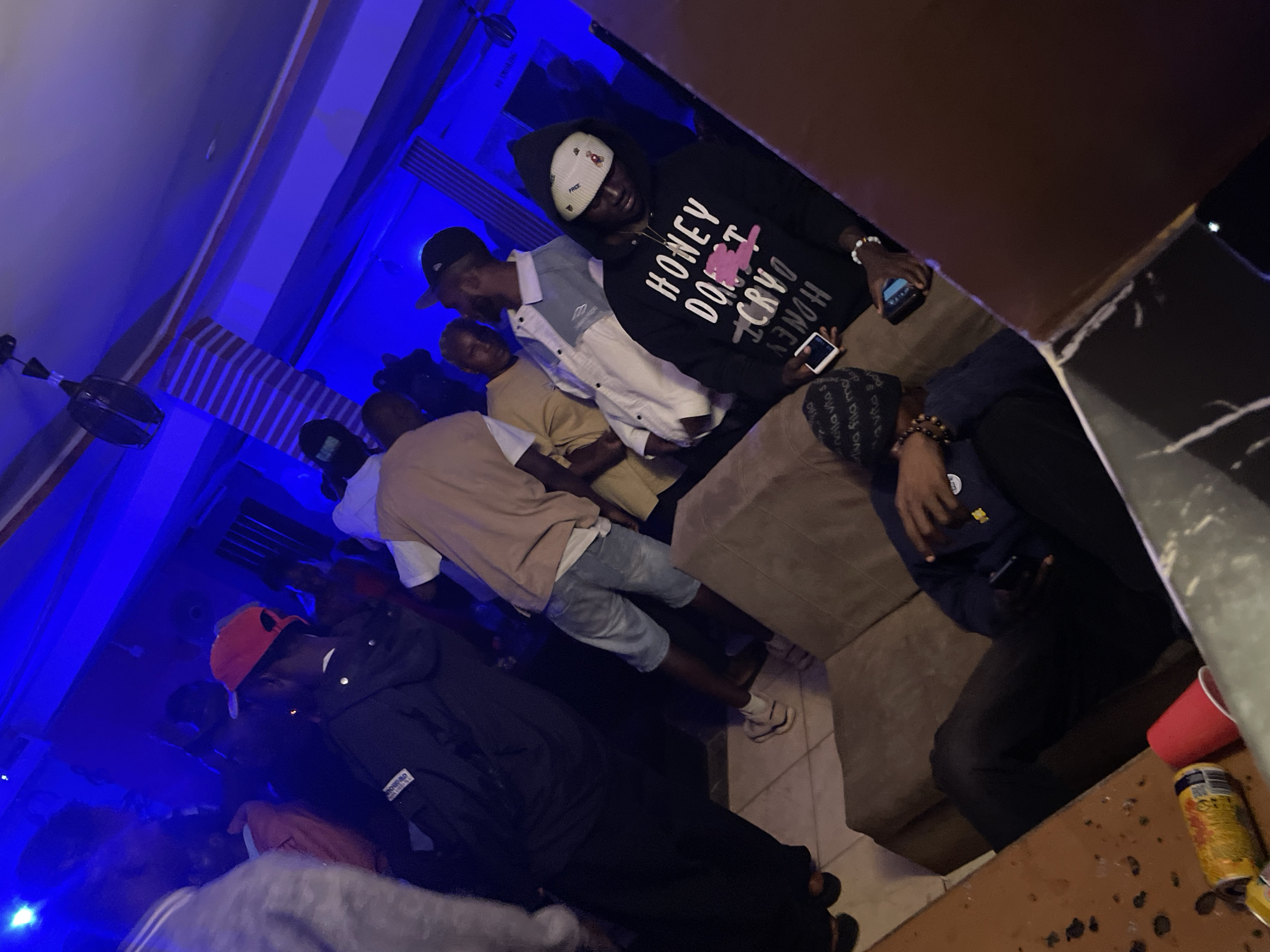 The image depicts a dimly lit indoor gathering, likely a party or casual get-together, with predominantly blue ambient lighting. In the foreground, a person in a black hoodie with white lettering that reads "HONEY DON'T CRY" is seated on a beige sofa, holding a mobile phone. To their left, another person is also seated, with their legs crossed and holding a mobile phone. On a nearby table, a red cup and two beverage cans are visible.
Further back, several other individuals are present. One person, wearing a red cap and a black jacket, stands near the center of the frame, facing away from the camera. Another individual is wearing a light-colored jacket over a white shirt and blue denim shorts. Other figures are blurred and partially visible in the background, suggesting a larger group. The environment appears to be a residential space or a small venue, with a patterned ceiling and walls. The overall atmosphere suggests a relaxed and social event. The provided information states the location is Durimi, Nigeria.