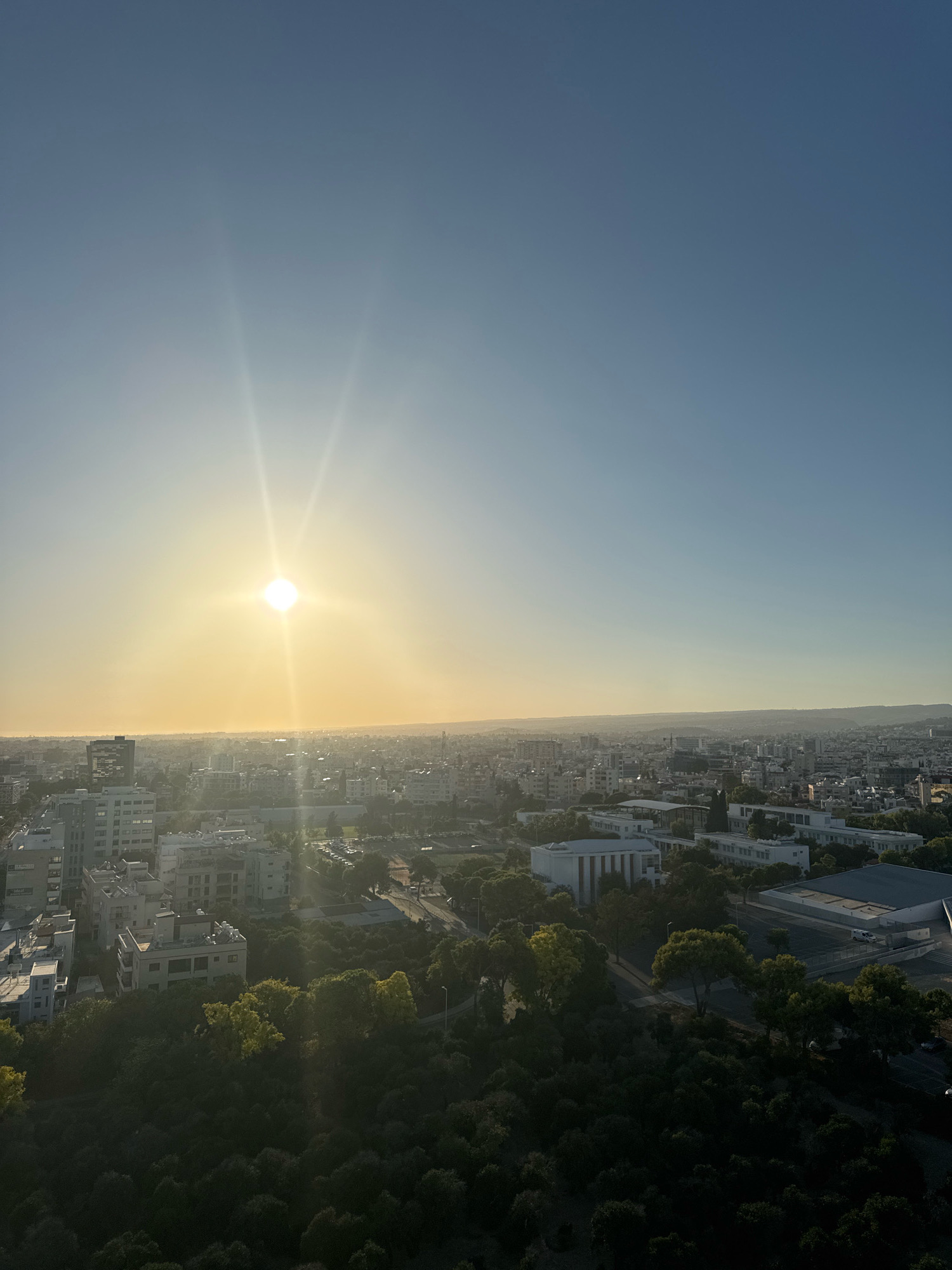 An aerial view of a city skyline in Limassol, Cyprus, at sunset. The sun is low on the horizon, casting a warm golden light across the scene and creating lens flares in the sky. A dense canopy of dark green trees dominates the foreground, with some lighter green foliage interspersed. Beyond the trees, the city unfolds with a variety of buildings, ranging from multi-story apartments to larger, modern structures with flat roofs. A few roads can be seen weaving through the urban landscape, with parked cars visible in some areas. The overall impression is one of a sprawling city bathed in the soft, ethereal glow of twilight.